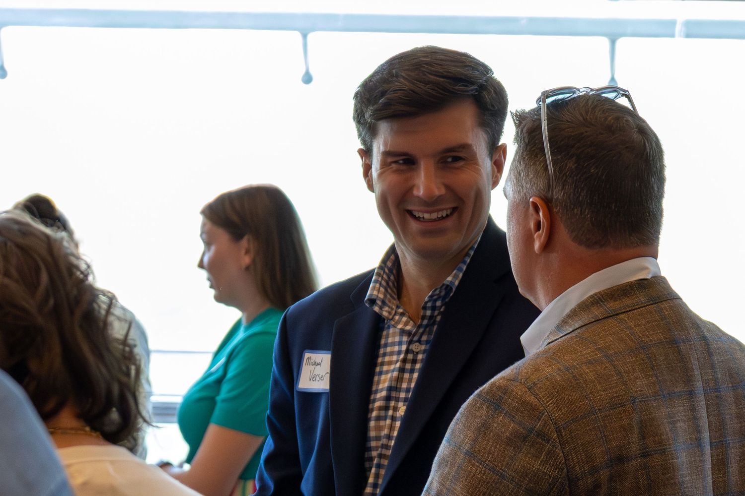 Professional networking event with smiling man in plaid shirt and blazer conversing with colleagues indoors.