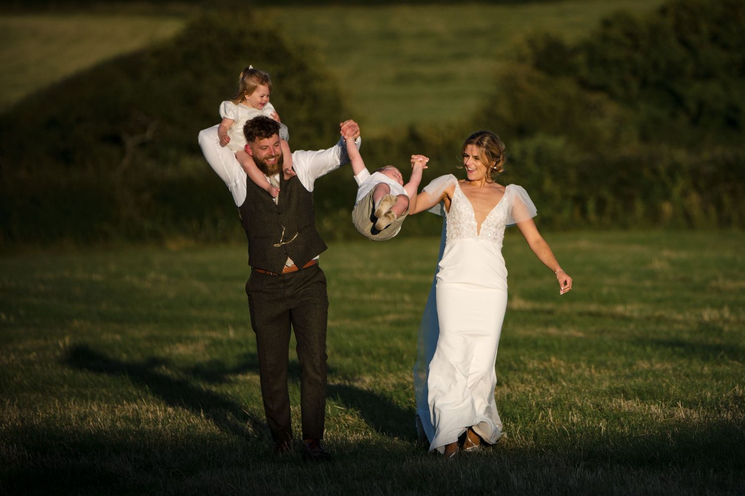 Bride and groom in wedding attire swinging a child between them in a sunny green field.