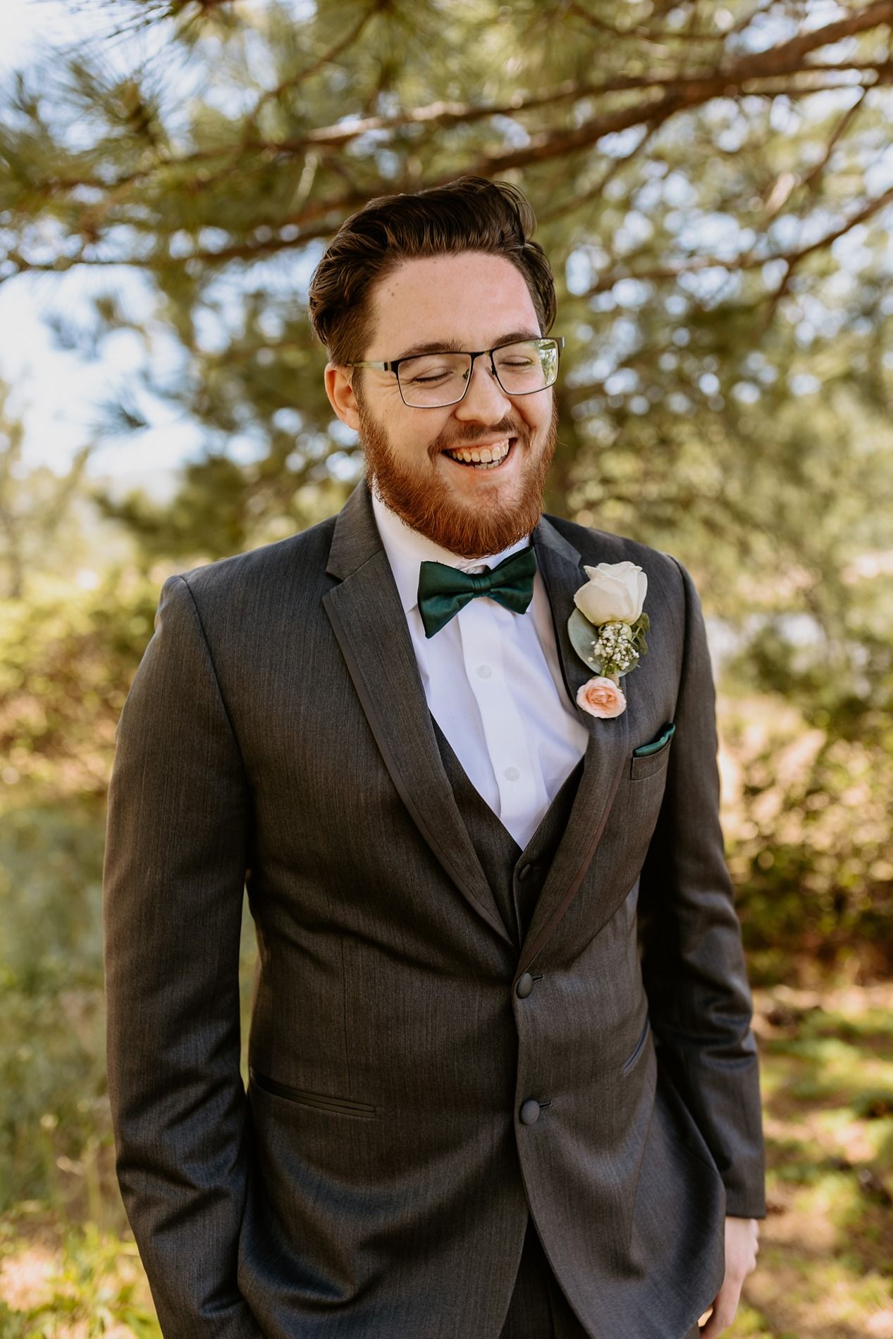 Smiling groom with red beard wearing gray suit, green bow tie, and white boutonniere outdoors at wedding at Lake Cuyamaca, Julian.