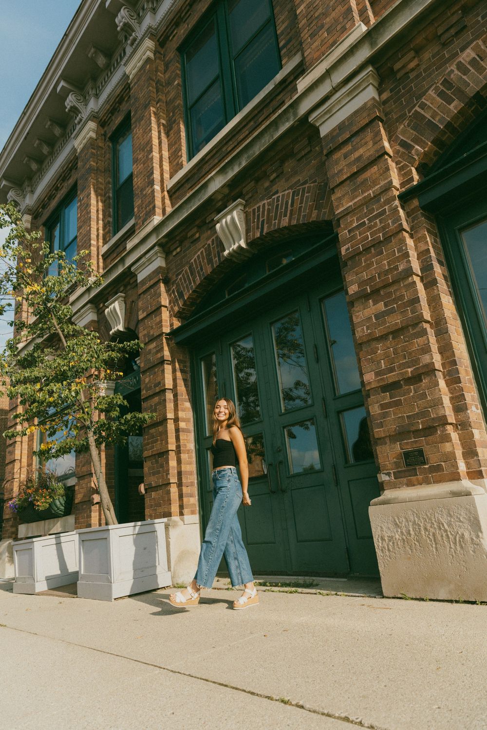 Woman in black top and wide-leg jeans walking outside a historic brick building with green doors on a sunny day.