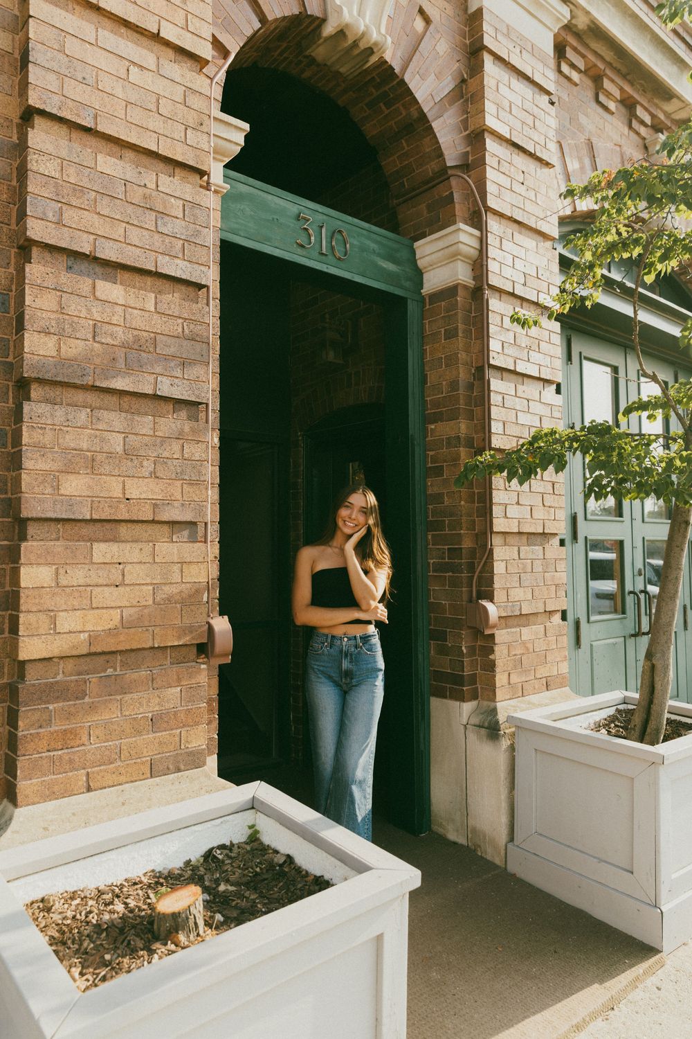 Young woman in black crop top and jeans standing in green arched doorway of brick building numbered 310.