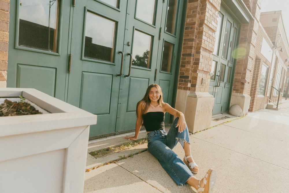 Young woman smiling, sitting on steps by a teal door, wearing black top and jeans, in an urban setting.