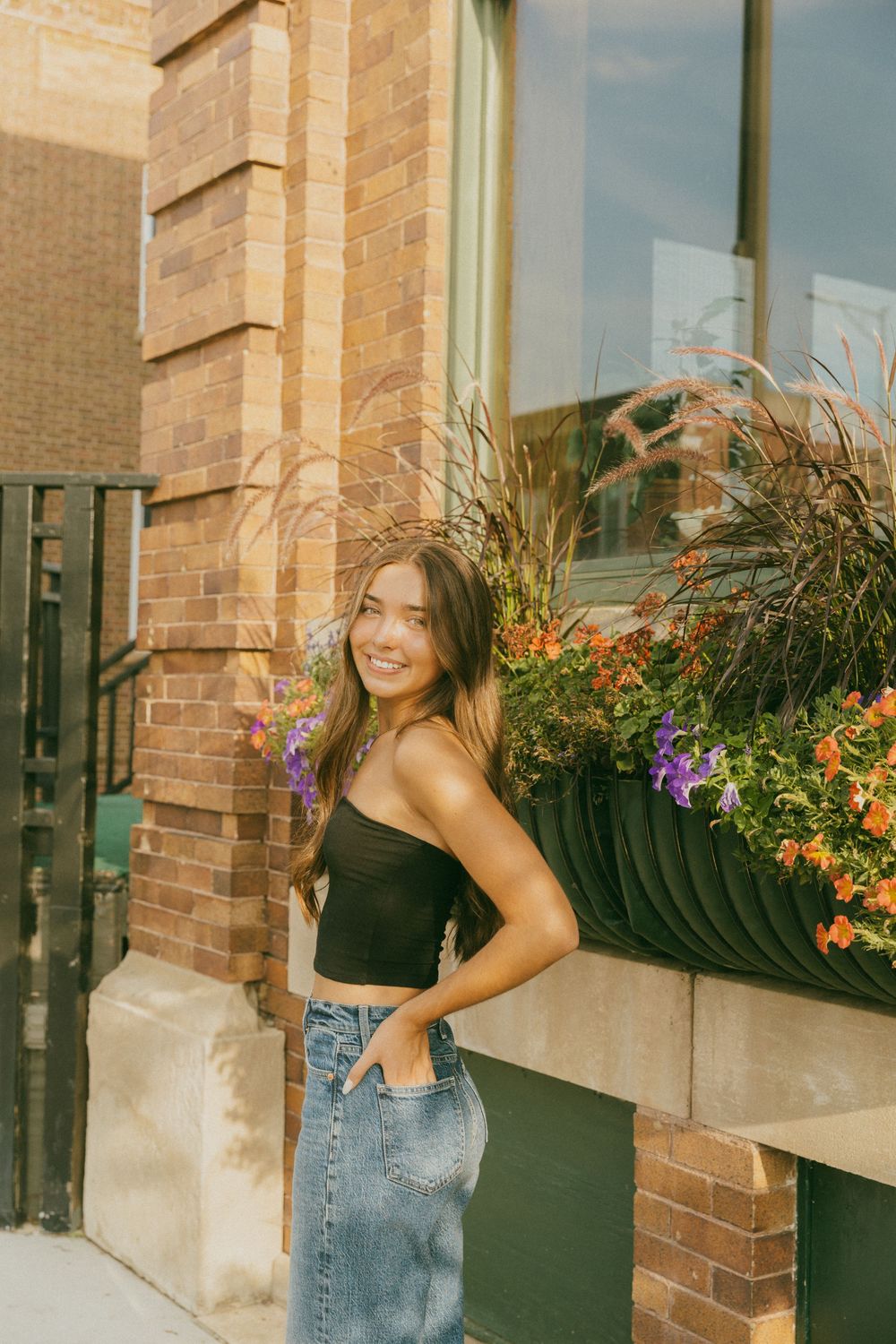 Smiling young woman in black crop top and denim skirt posing beside colorful flower box on urban brick building sidewalk.