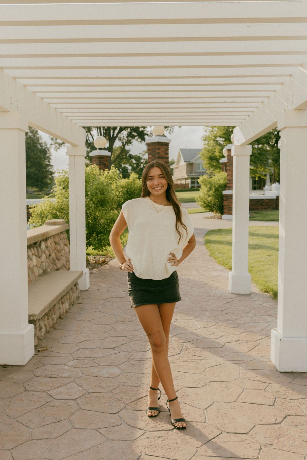 Smiling young woman in white top and dark mini skirt posing under white pergola in lush garden setting.
