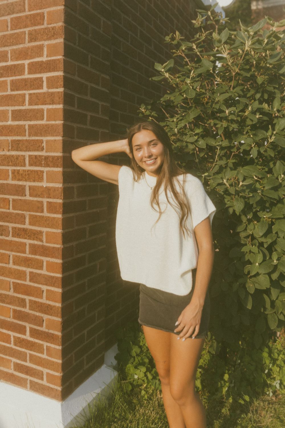 Smiling young woman in white tee and dark shorts posing by brick wall with green shrubs outdoors.