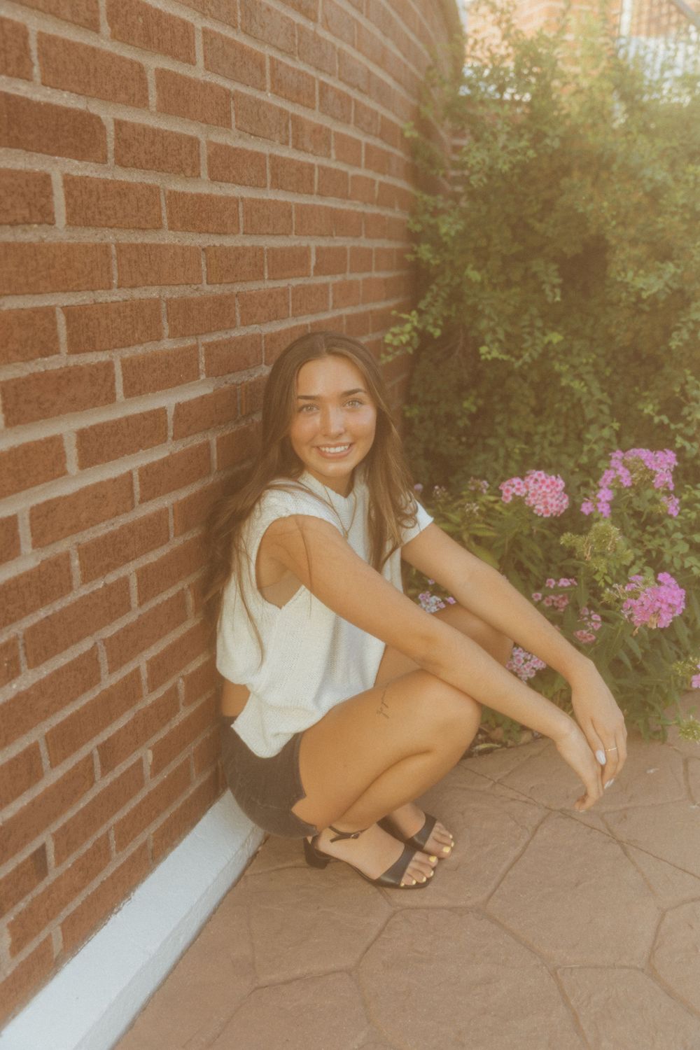 Young woman smiling and crouching against a red brick wall with pink flowers and greenery in the background.