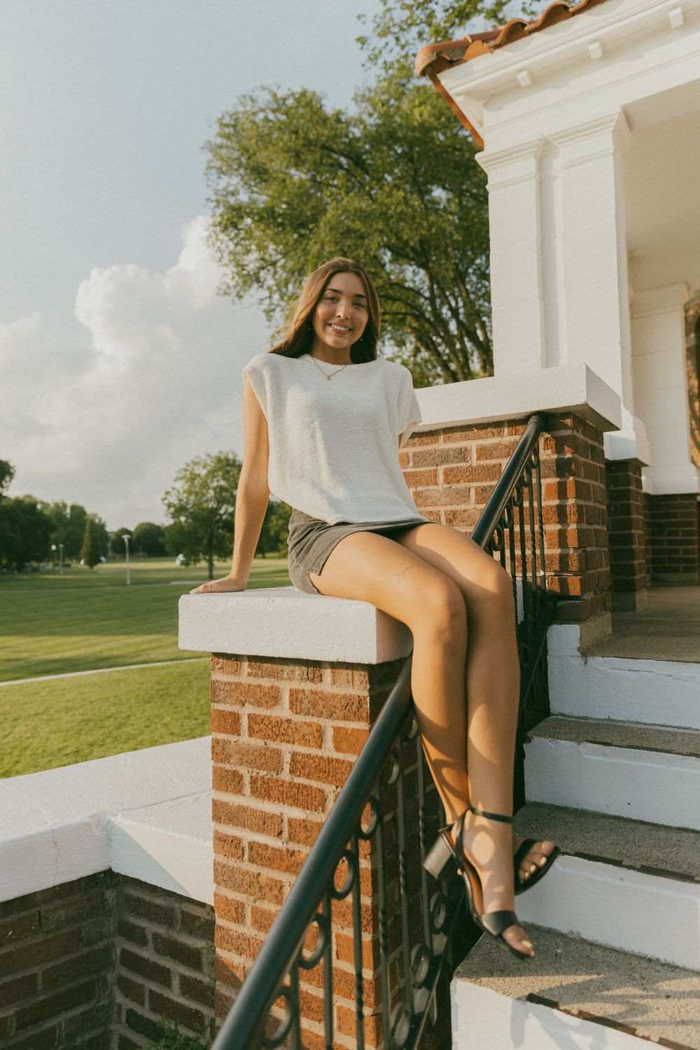 Young woman in white top and dark mini skirt sitting on brick staircase railing outside white building on sunny day.