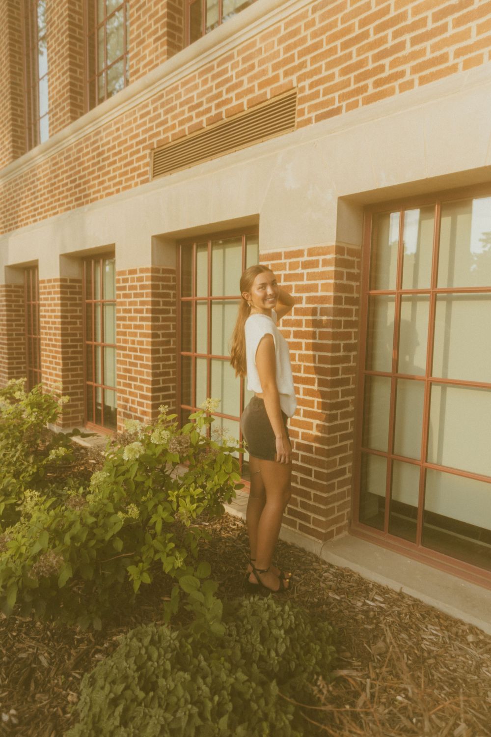 Young woman in white top and black shorts smiling beside a brick building with large windows and green shrubs.