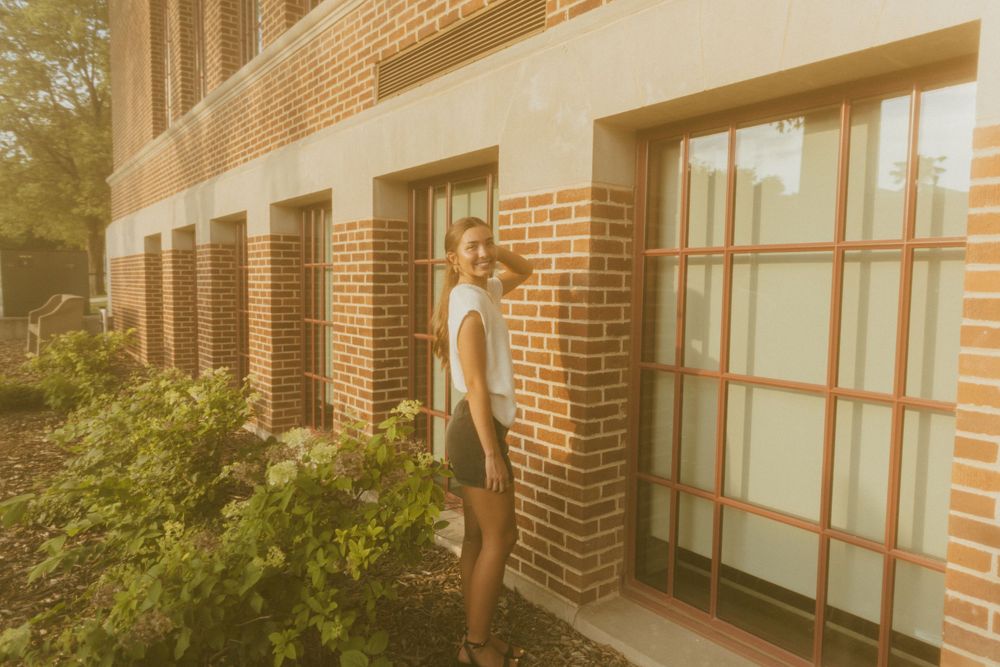 Young woman posing beside a brick building with large windows, surrounded by lush green plants in warm golden light.