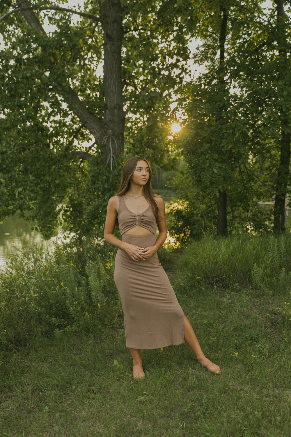 Young woman in tan two-piece midi dress standing barefoot in lush green forest at golden sunset by water.