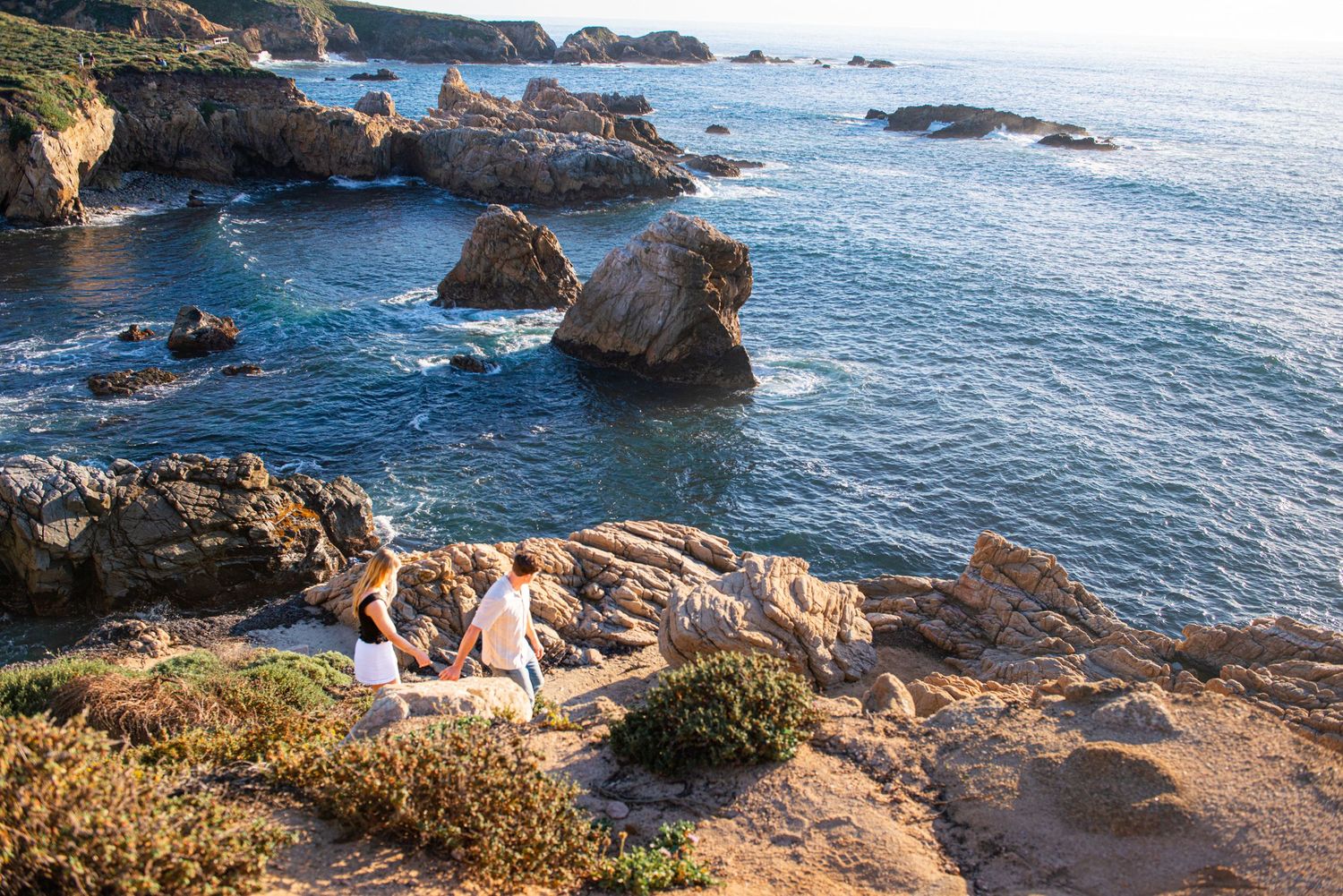 Couple embracing on a scenic coastal trail with rocky cliffs and blue ocean waves at Big Sur, California.