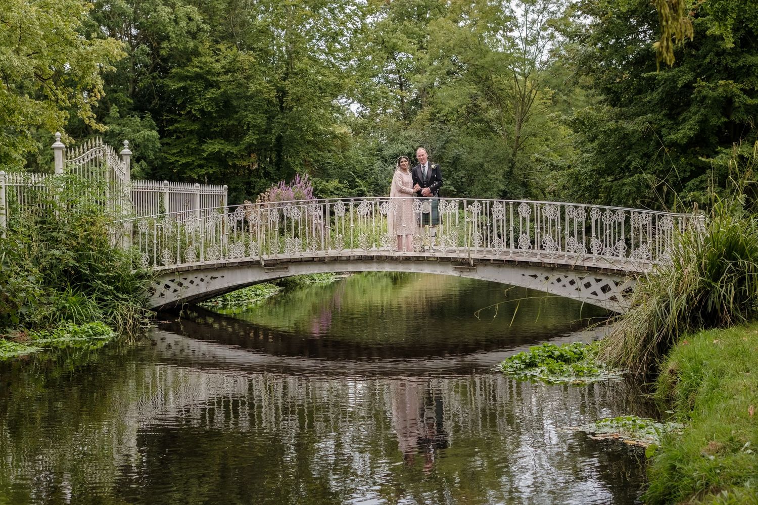Couple embracing on ornate white bridge over calm pond in lush green garden setting.