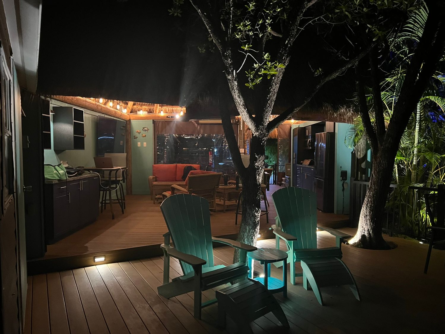 A cozy outdoor deck at night features Adirondack chairs, string lights, and tropical trees surrounding a teal accent table.