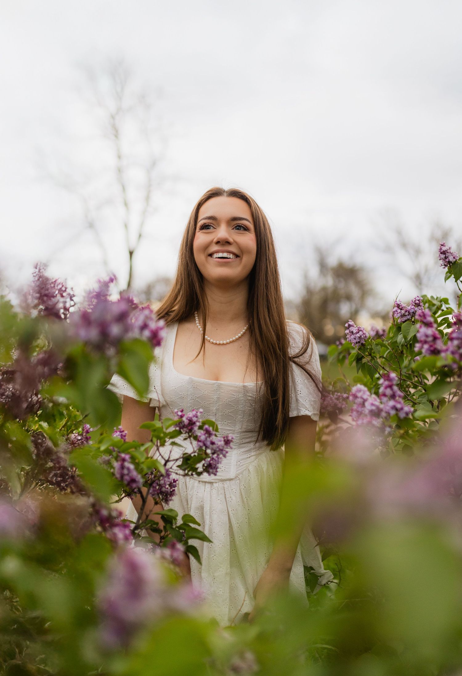 Young woman in white dress smiling among blooming purple lilac bushes in a garden setting.