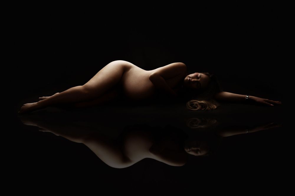 Artistic nude silhouette of a woman lying on a reflective surface against a dark background.