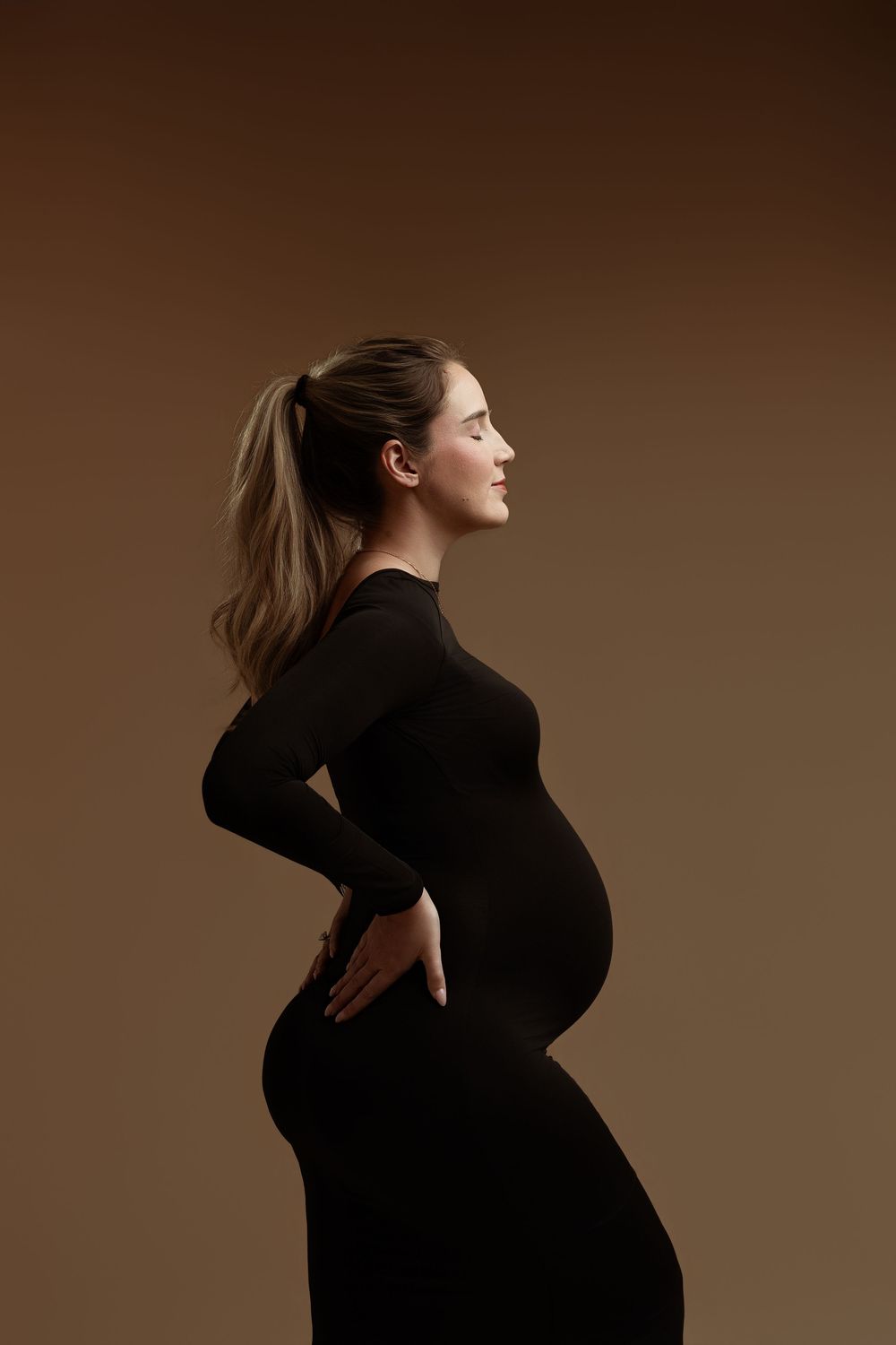 Pregnant woman in black dress holding her baby bump, side profile studio shot on brown background.