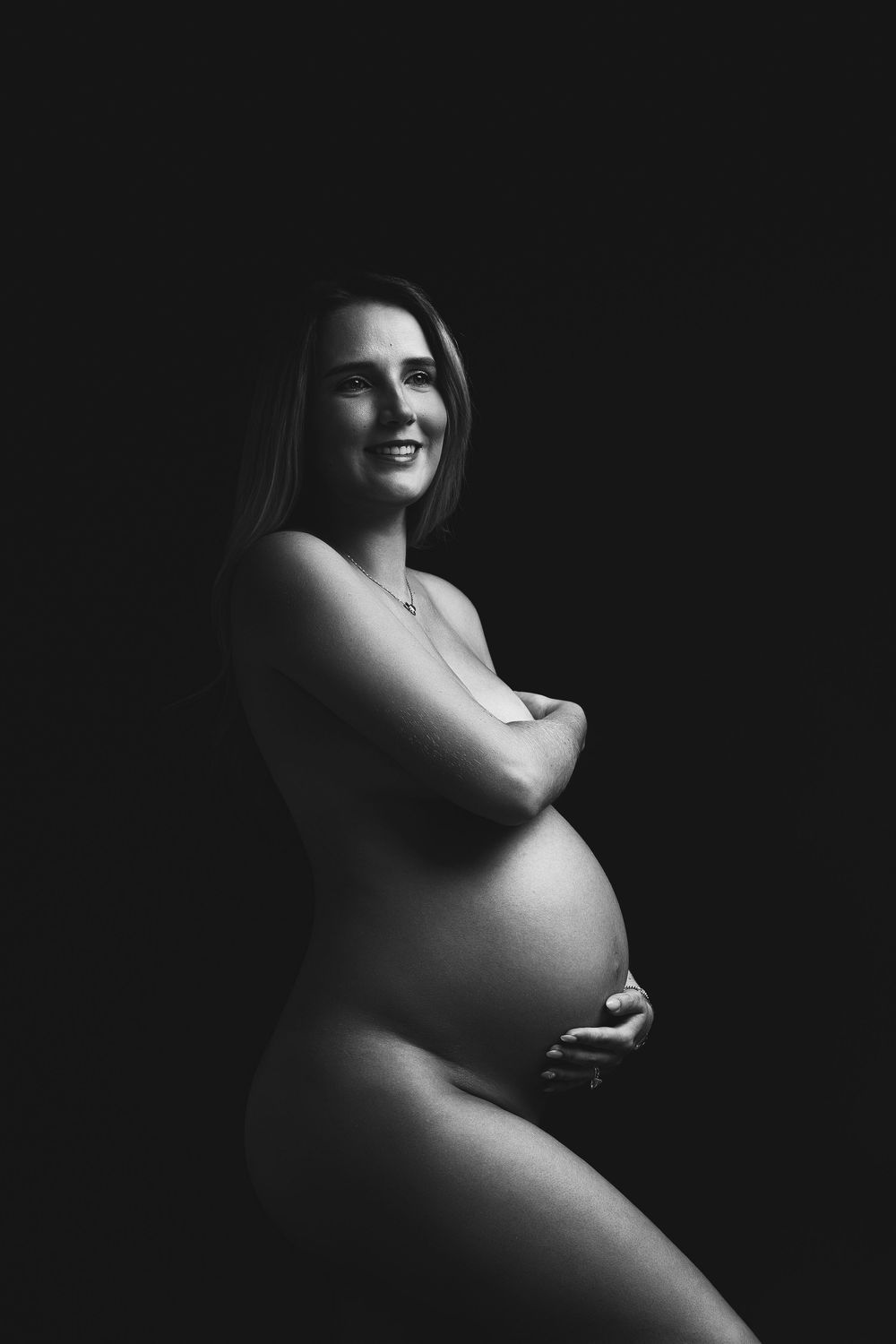 Black and white maternity portrait of smiling pregnant woman posing gracefully against dark background.