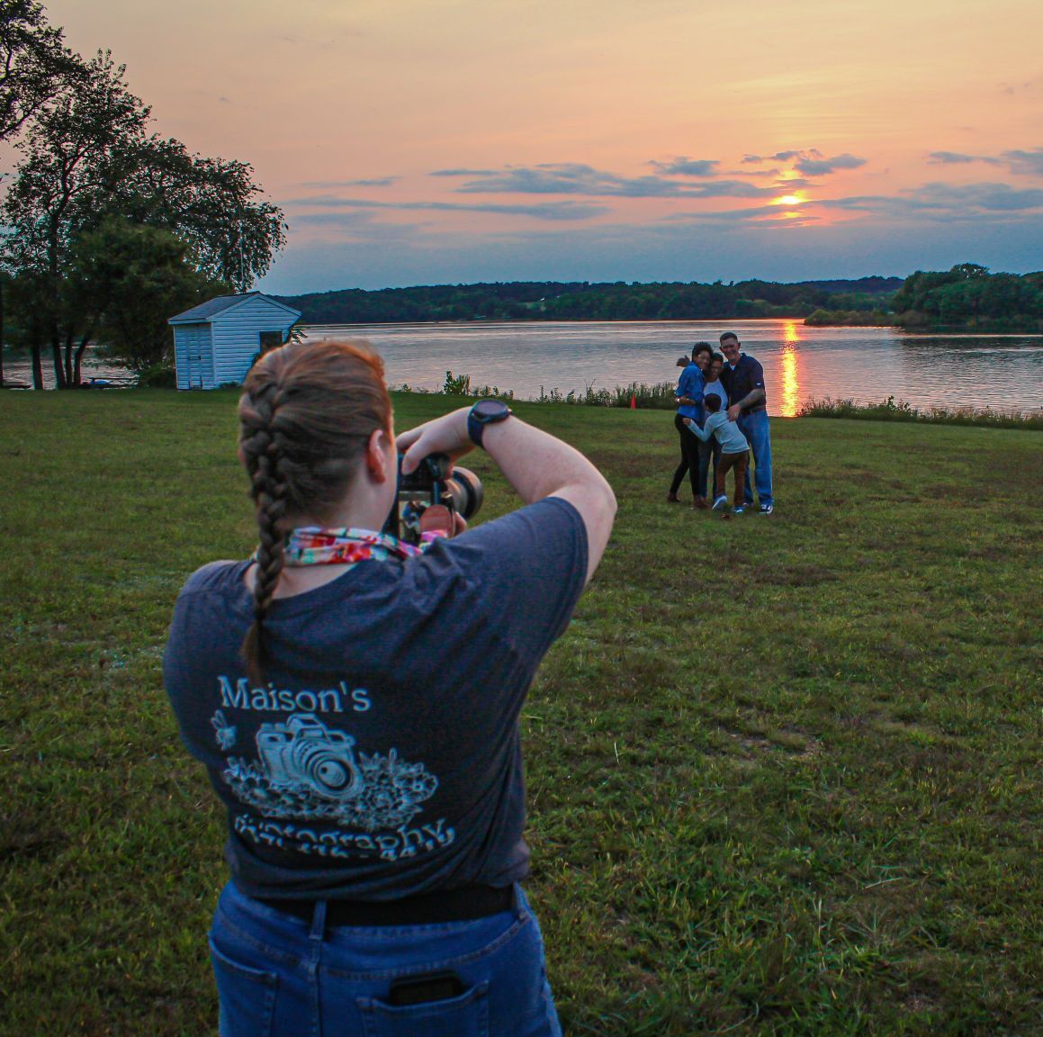 A photographer takes pictures of a couple posing by a lake during a vibrant sunset.
