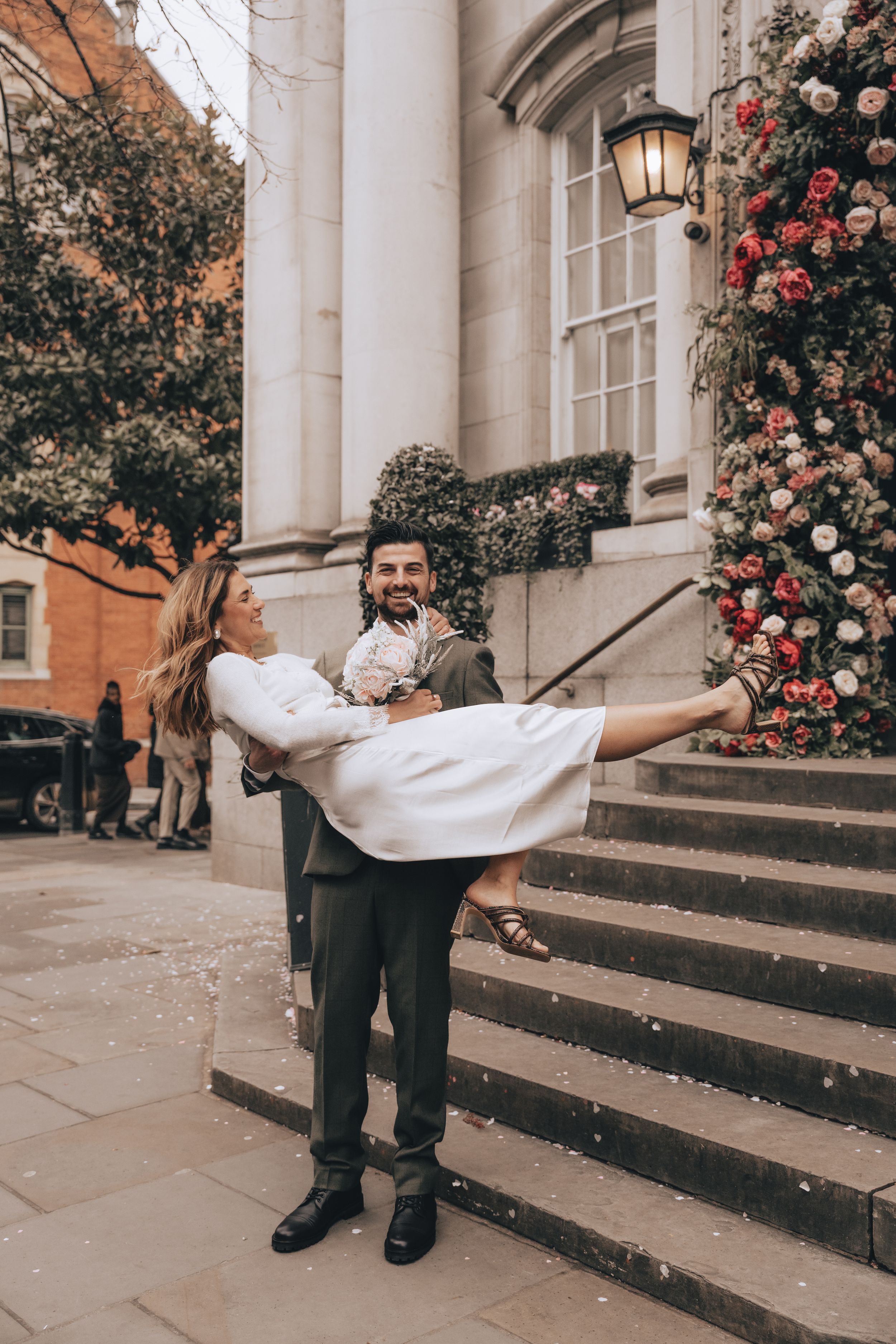 Groom carrying bride in white dress outside decorated Chelsea Old Town Hall wedding venue with floral arch and stone steps.