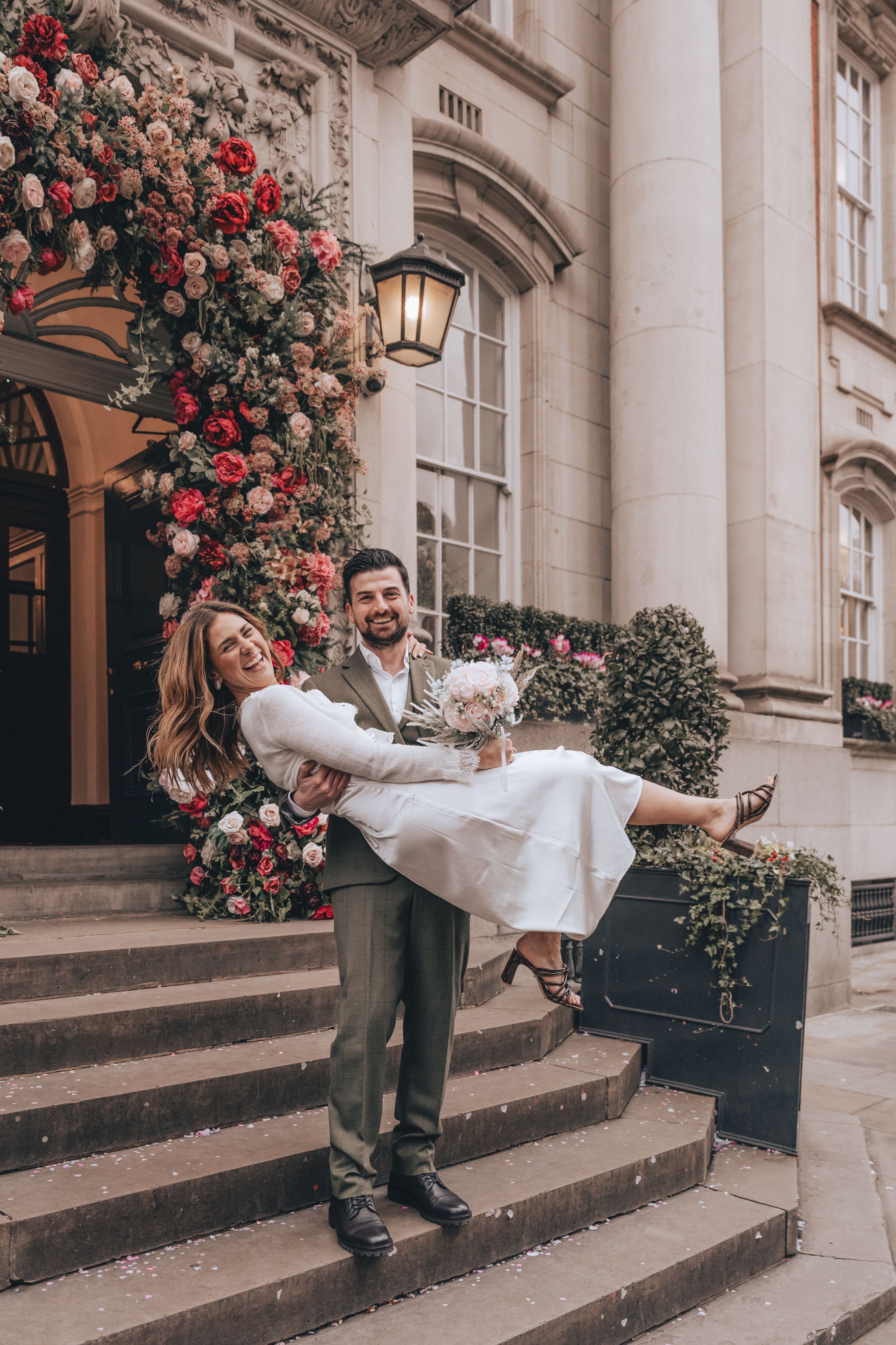 Joyful groom carrying bride on steps of the elegant building of Chelsea Old Town Hall adorned with lush floral arrangements.