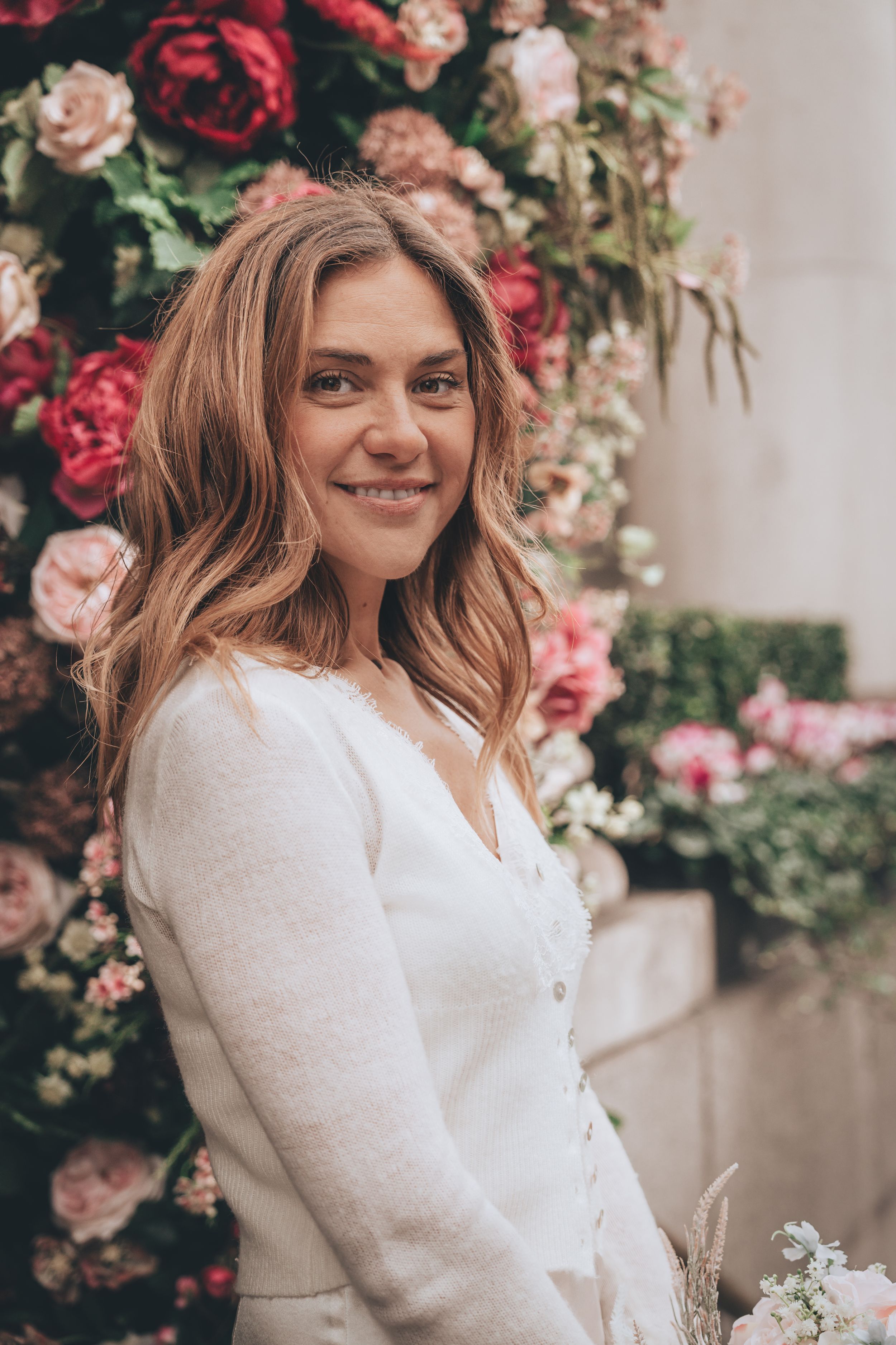 Smiling bride woman in white cardigan poses before a lush floral backdrop of pink and red roses at Chelsea Old Town Hall entrance.