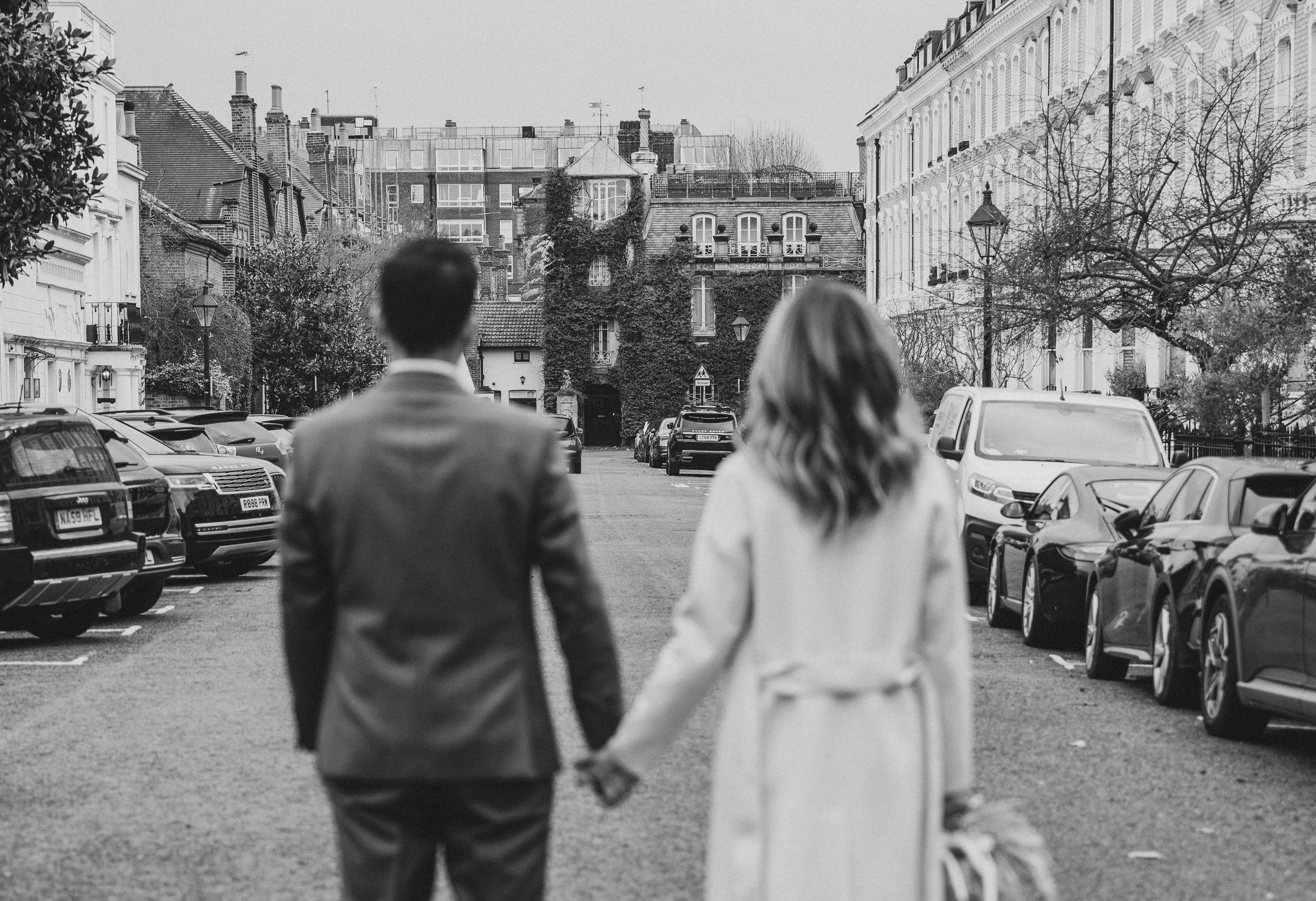 Black and white photo of a couple holding hands walking down a city street lined with parked cars.