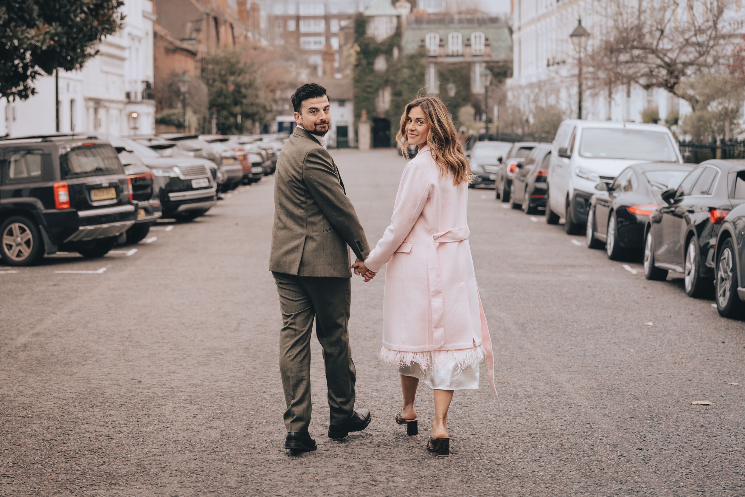 Couple holding hands walking down a quiet street, woman in pink dress and man in olive suit, urban setting.