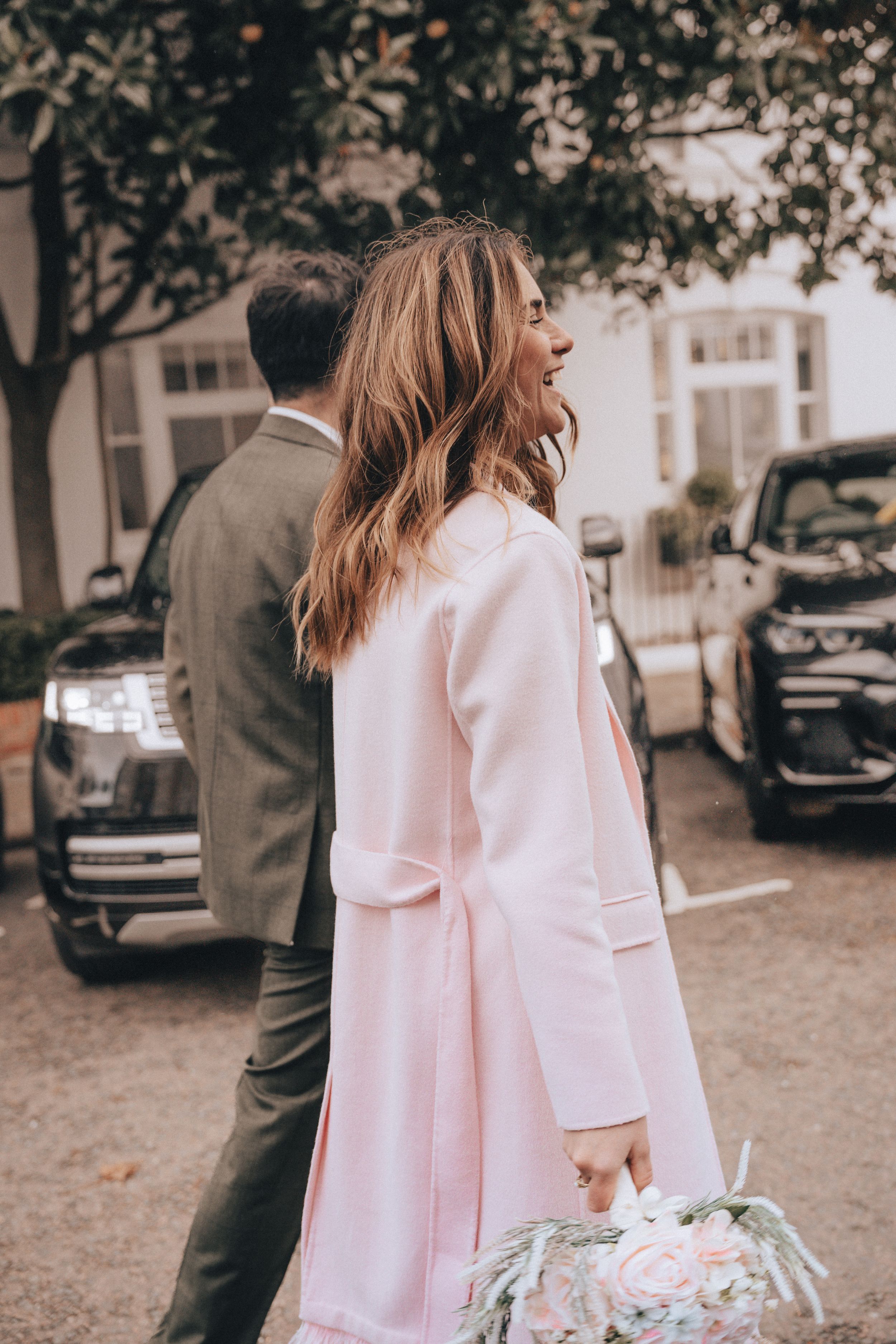 Woman in pink coat holding bouquet walks with man in olive suit near parked cars outdoors.