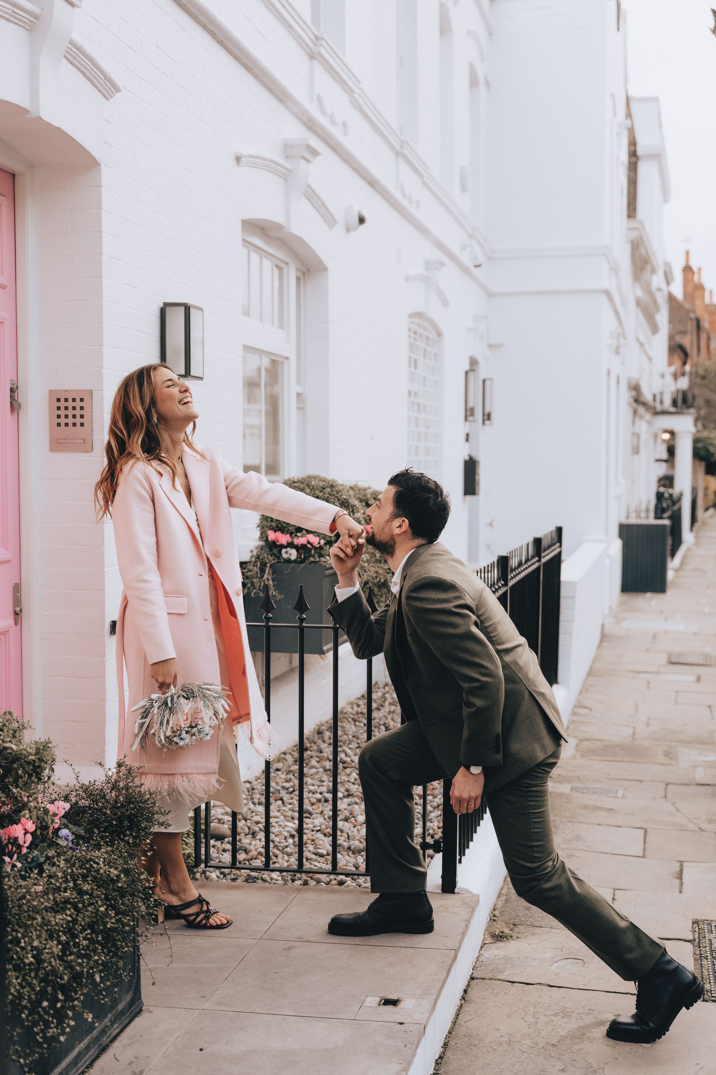 Man kneeling to kiss woman's hand on a charming white London street, both dressed in elegant outfits.