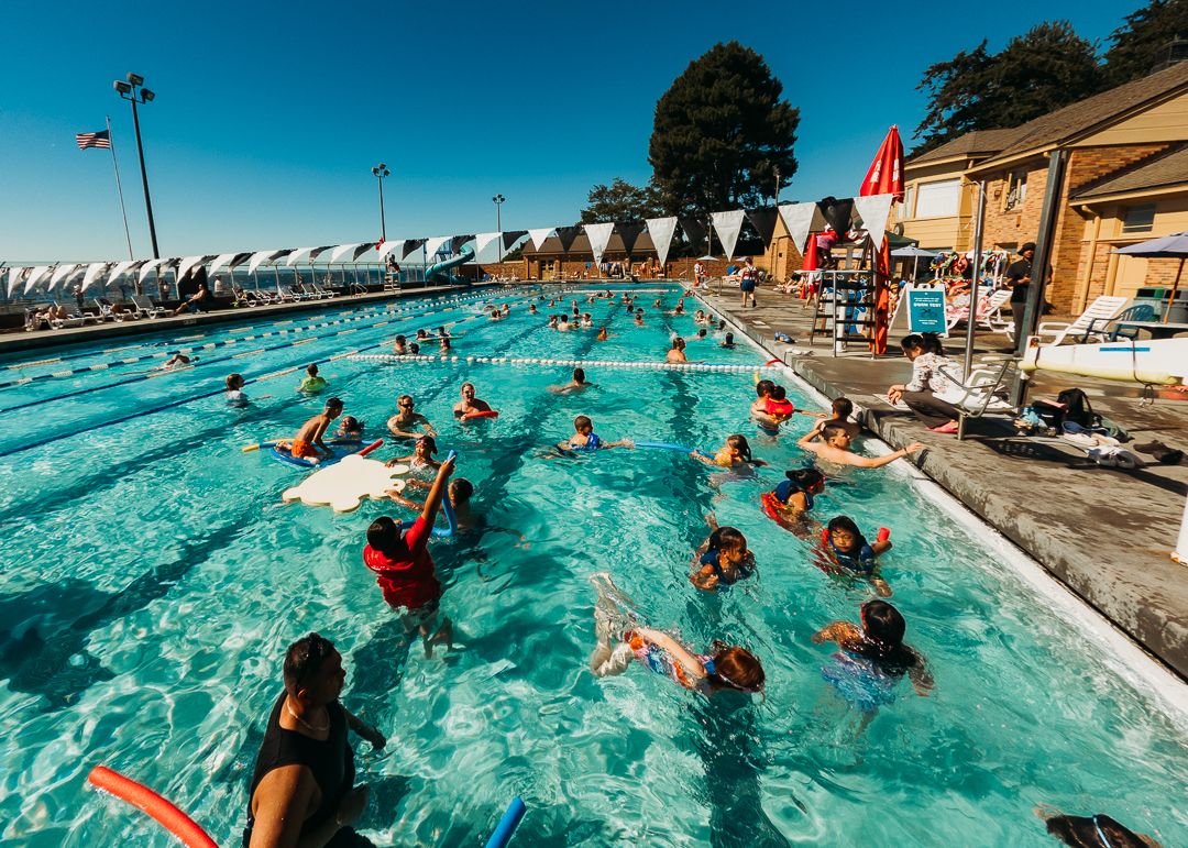 Crowded outdoor swimming pool with swimmers enjoying a sunny day, surrounded by lounge chairs and a pool house.