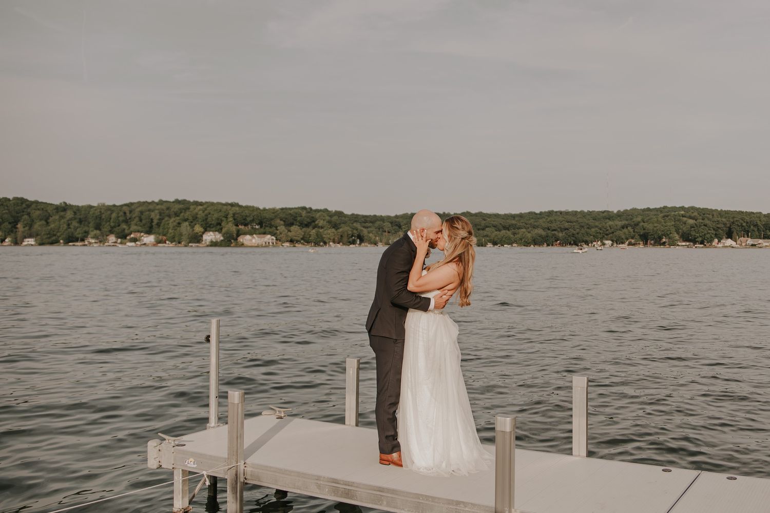 Bride and groom share a romantic kiss on a lakeside dock surrounded by serene water and lush green trees.