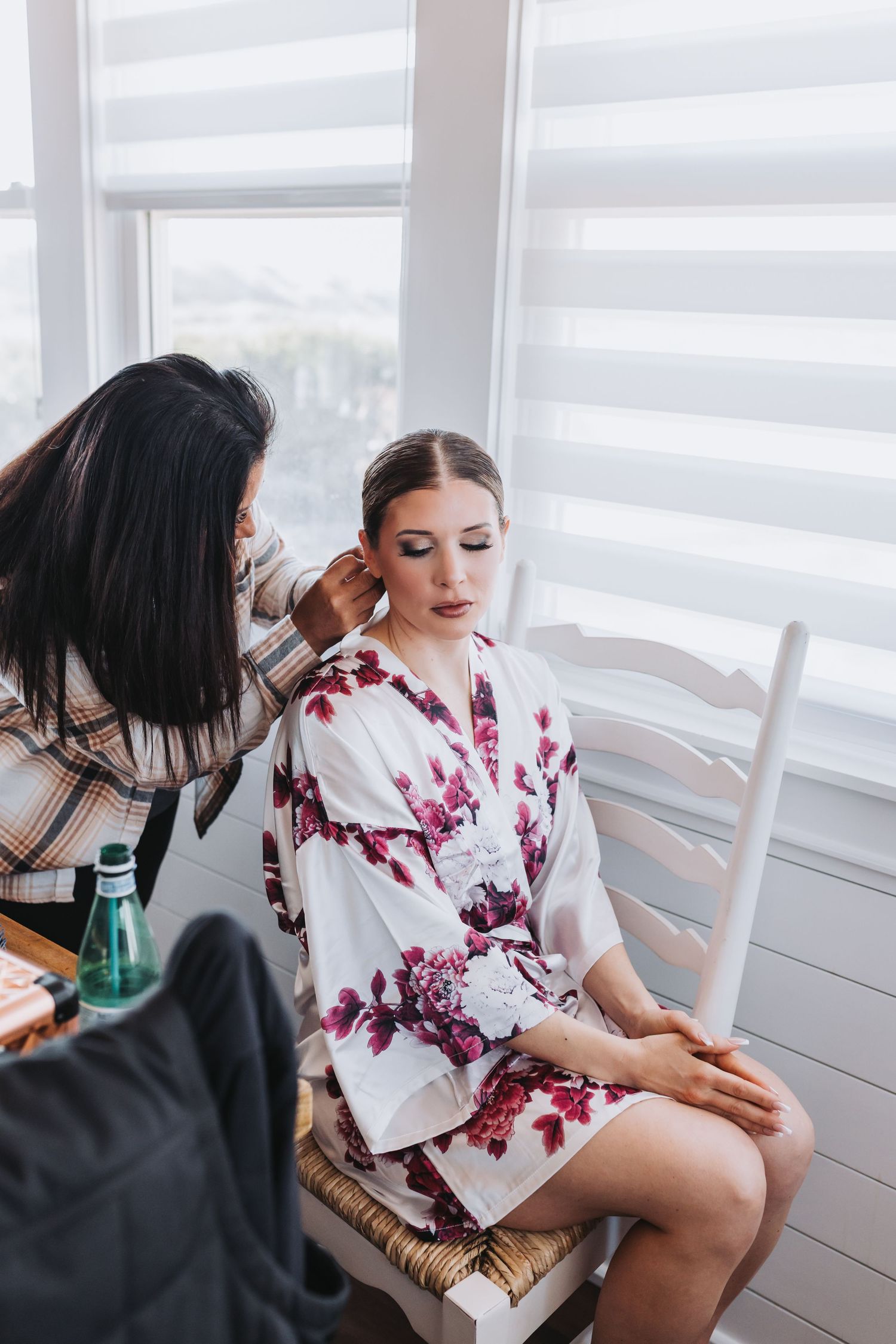 Bride in floral robe getting makeup done by artist near bright window before wedding ceremony.