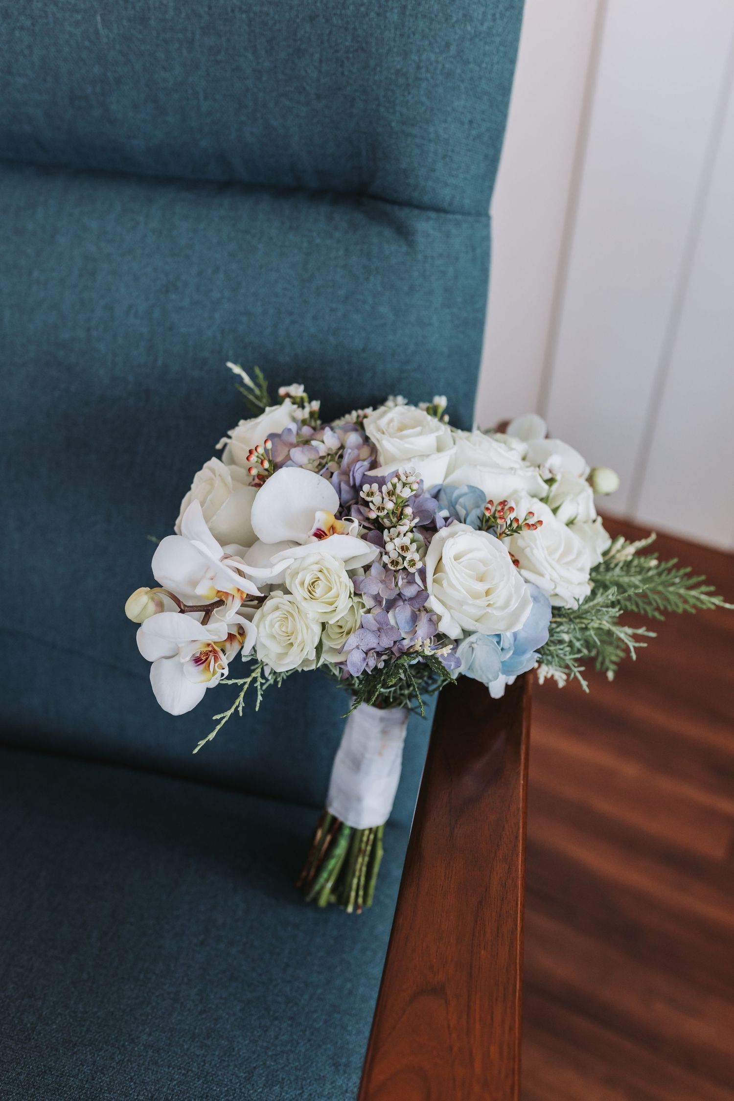 Elegant bridal bouquet with white roses, orchids, and blue hydrangeas resting on a teal velvet chair.