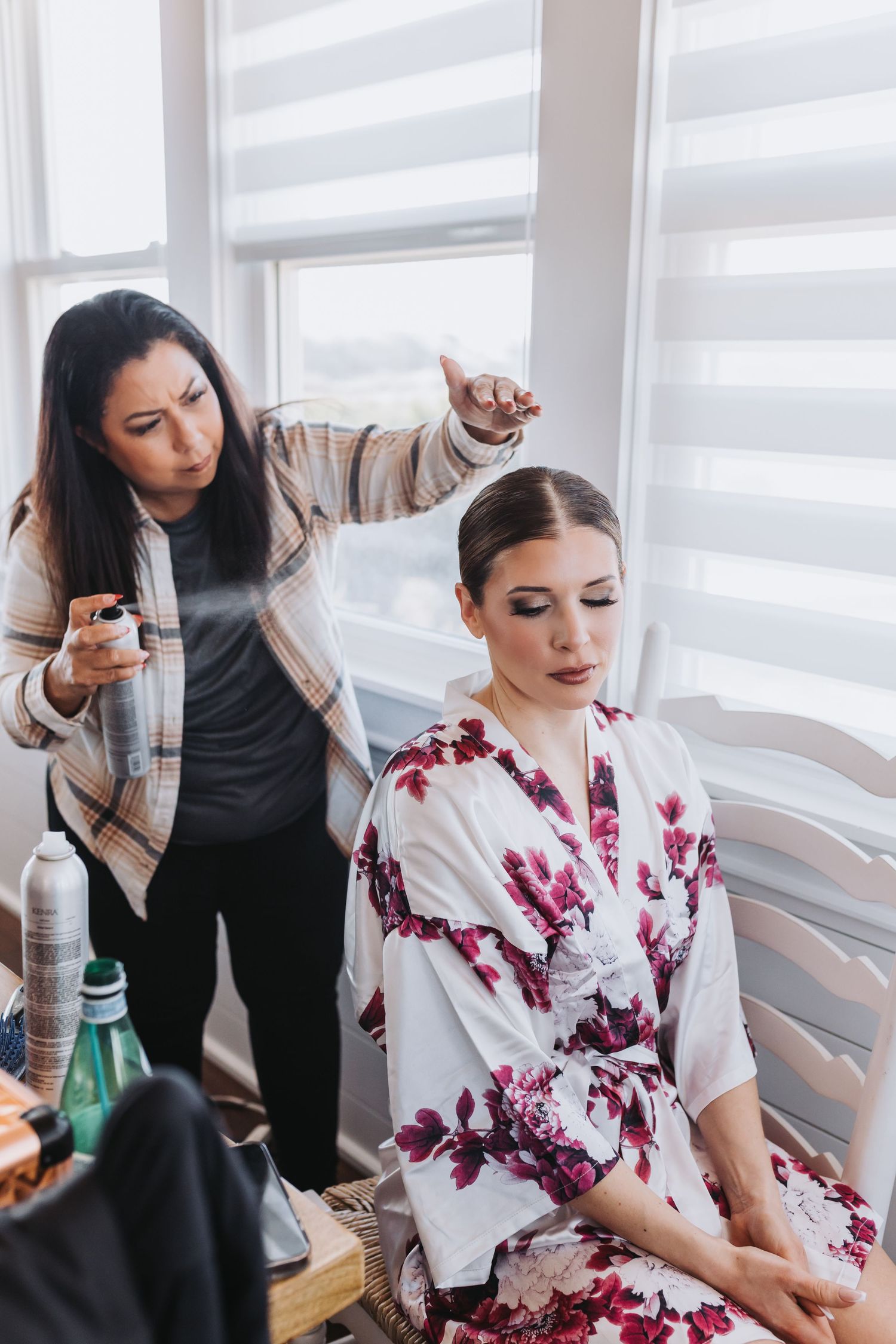 Hair stylist applying hairspray to bride's elegant updo while she sits in floral robe near bright windows.
