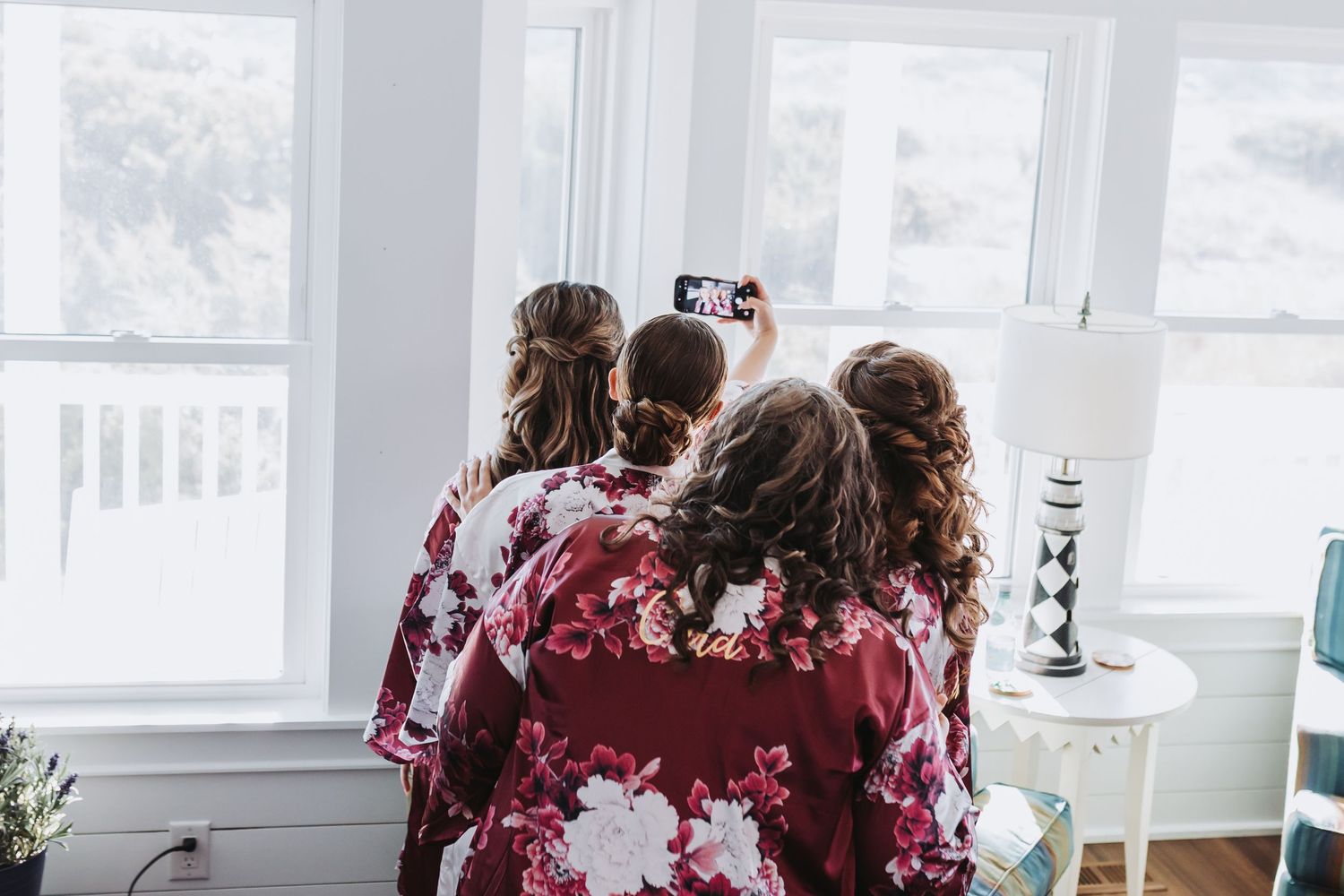 Three bridesmaids in matching floral robes taking a selfie together near a bright window.