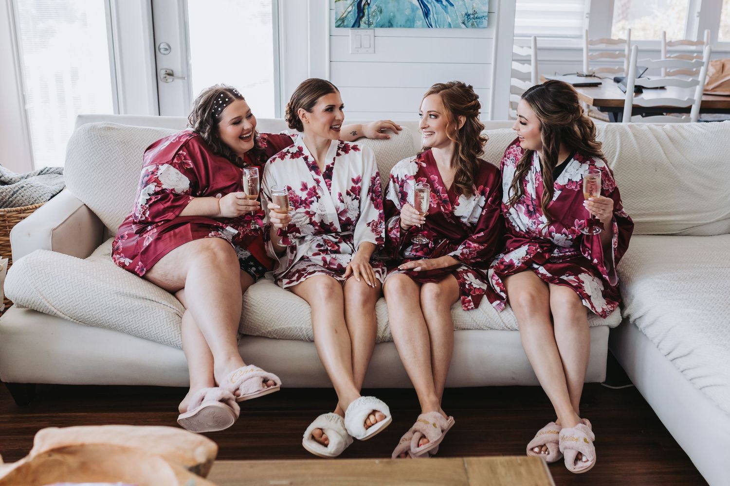 Four bridesmaids laughing together on white sofa wearing matching floral robes and fuzzy slippers.