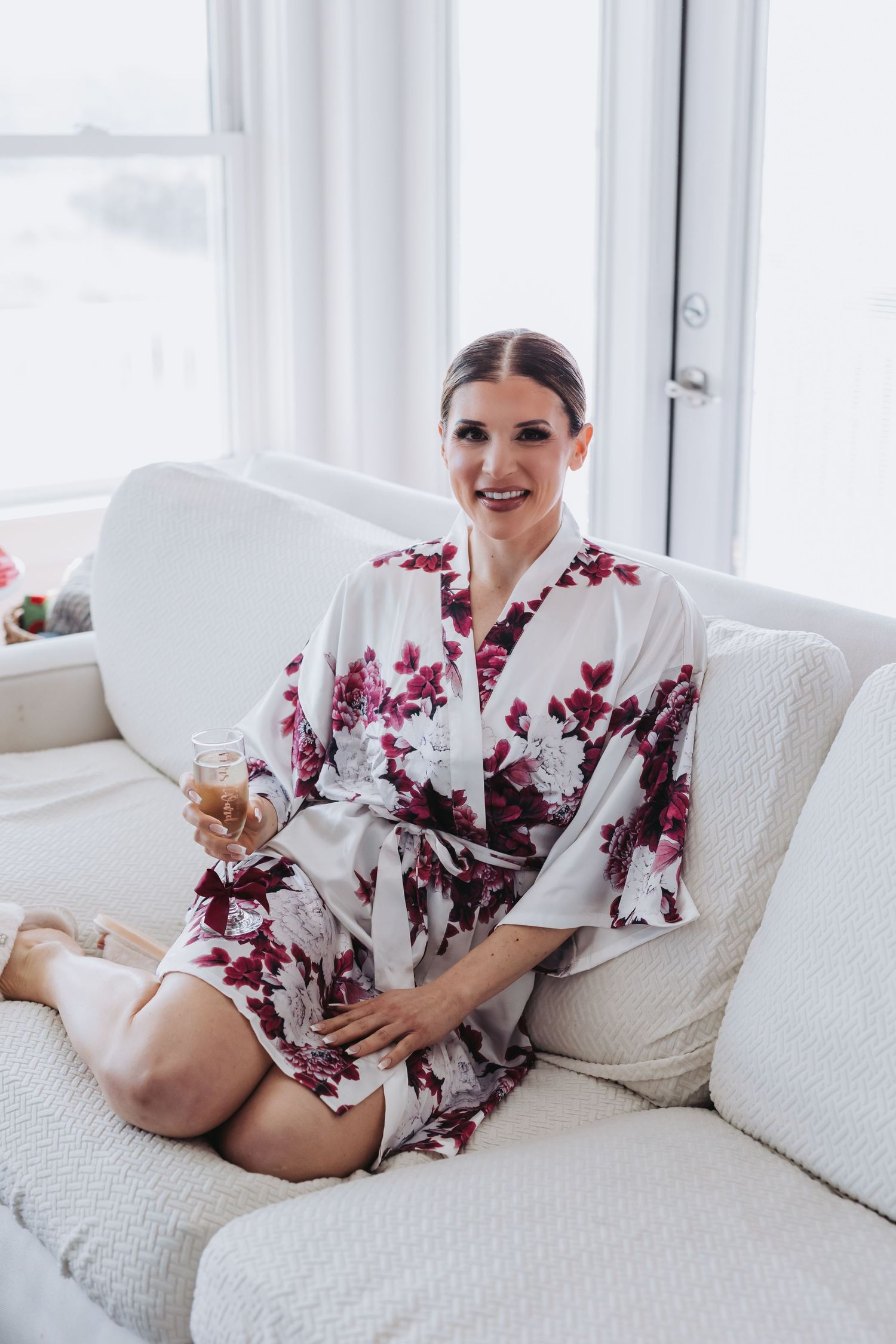Bride relaxes on white sofa in floral kimono robe holding champagne glass during wedding preparations.