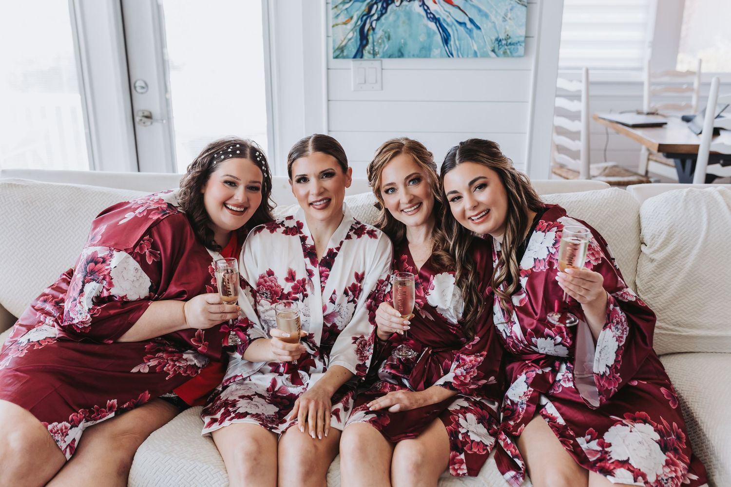 Four bridesmaids in matching burgundy floral robes toasting with champagne glasses on a white sofa.
