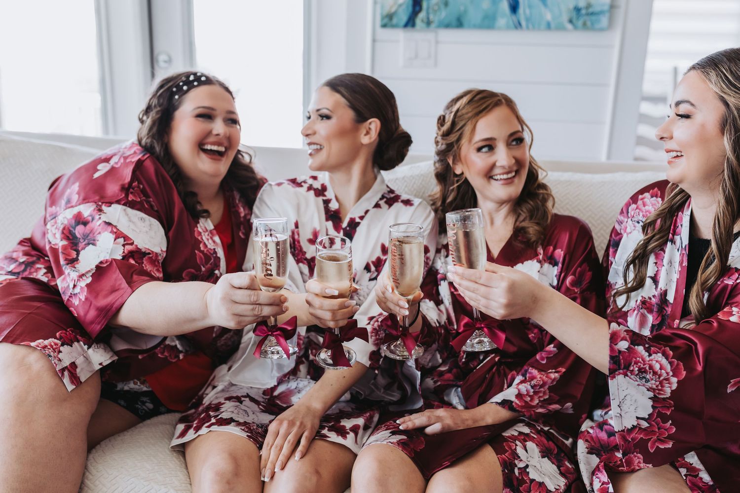 Four bridesmaids in floral robes toast with champagne glasses, laughing together before the wedding.