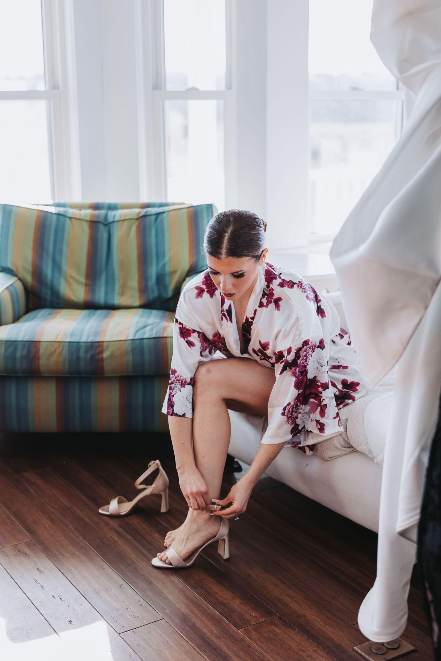 Bride in floral robe puts on heels while sitting on white chair near bright window.