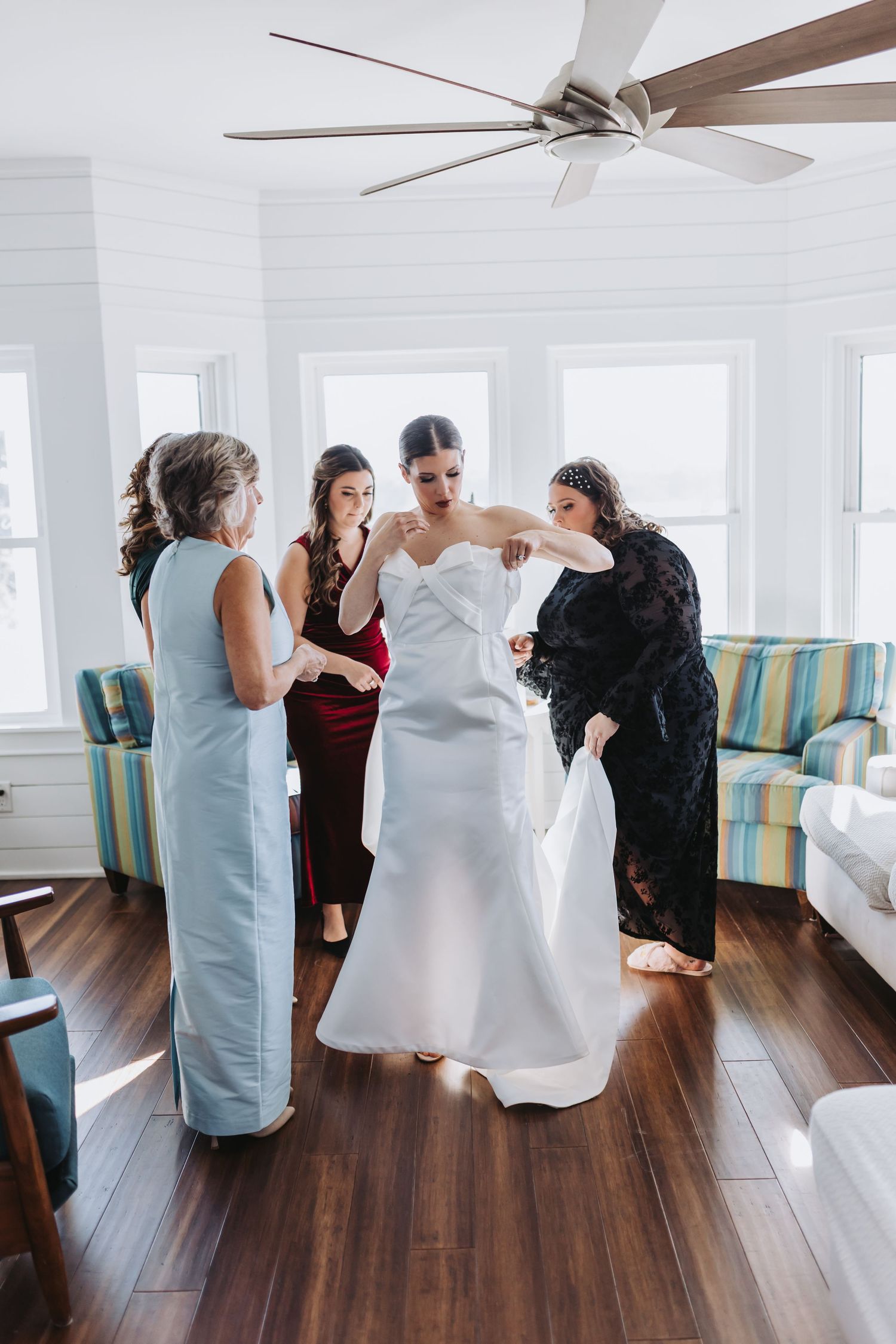 Bridesmaids helping bride into white wedding gown in bright coastal room with hardwood floors and ceiling fan.