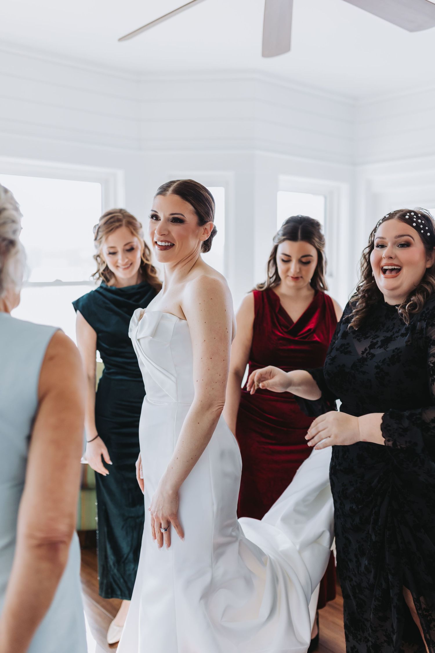 Excited bridesmaids in colorful dresses help bride into white wedding gown indoors before ceremony.