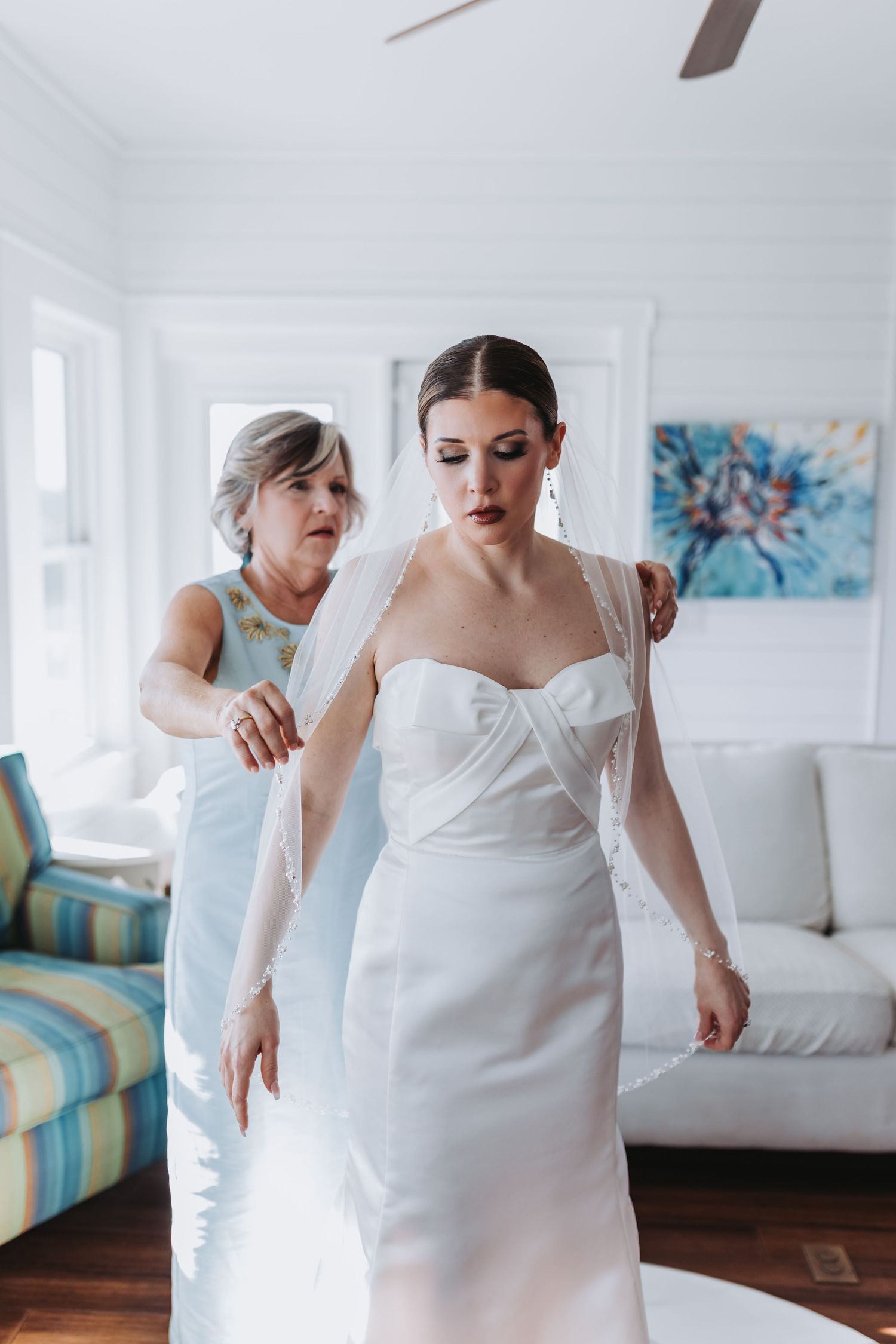 A bride in a white gown gets her veil adjusted by a woman in a blue dress in a bright white room.