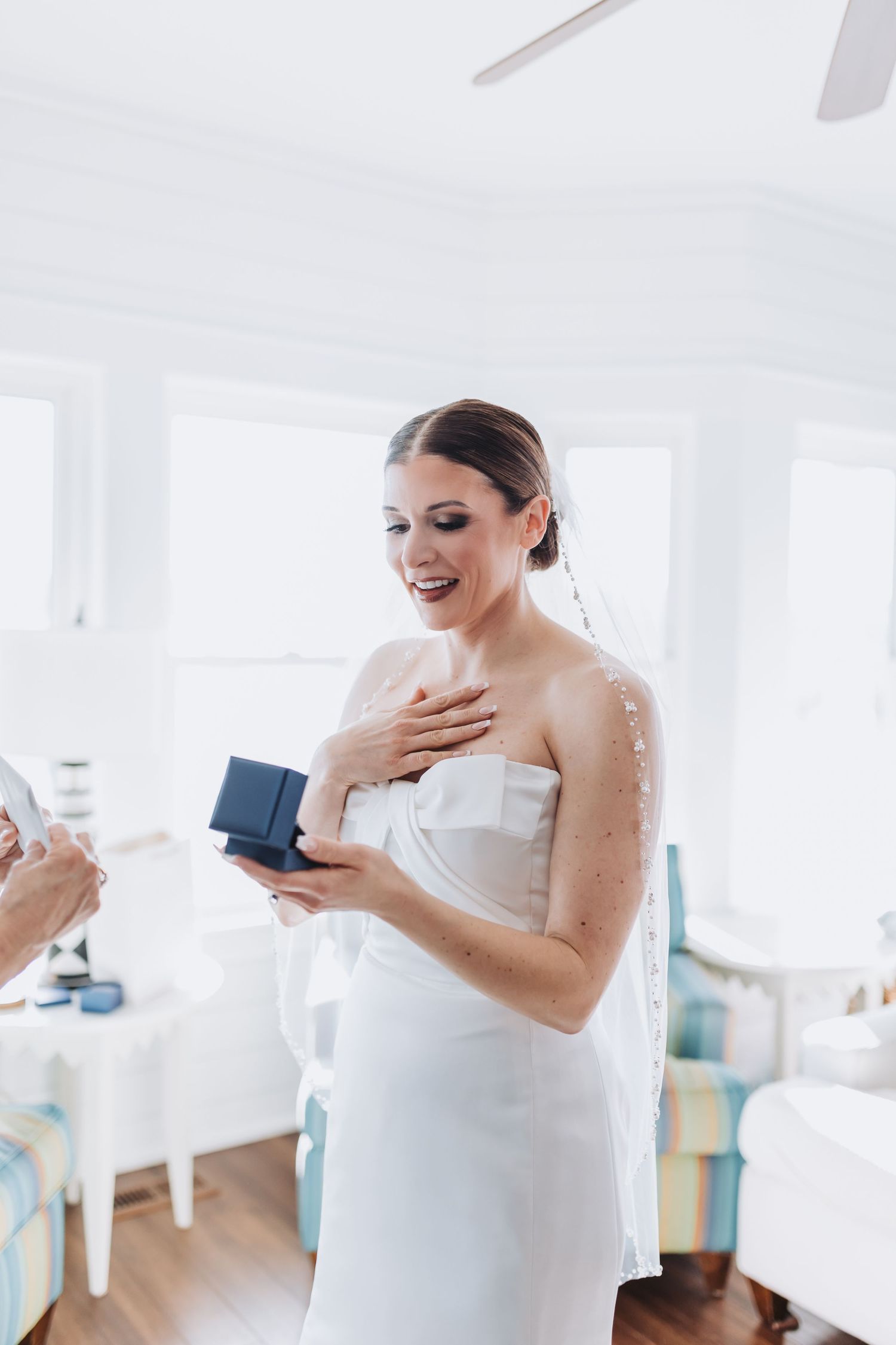 Bride in strapless white gown reacts with joy while receiving a gift box on her wedding day.