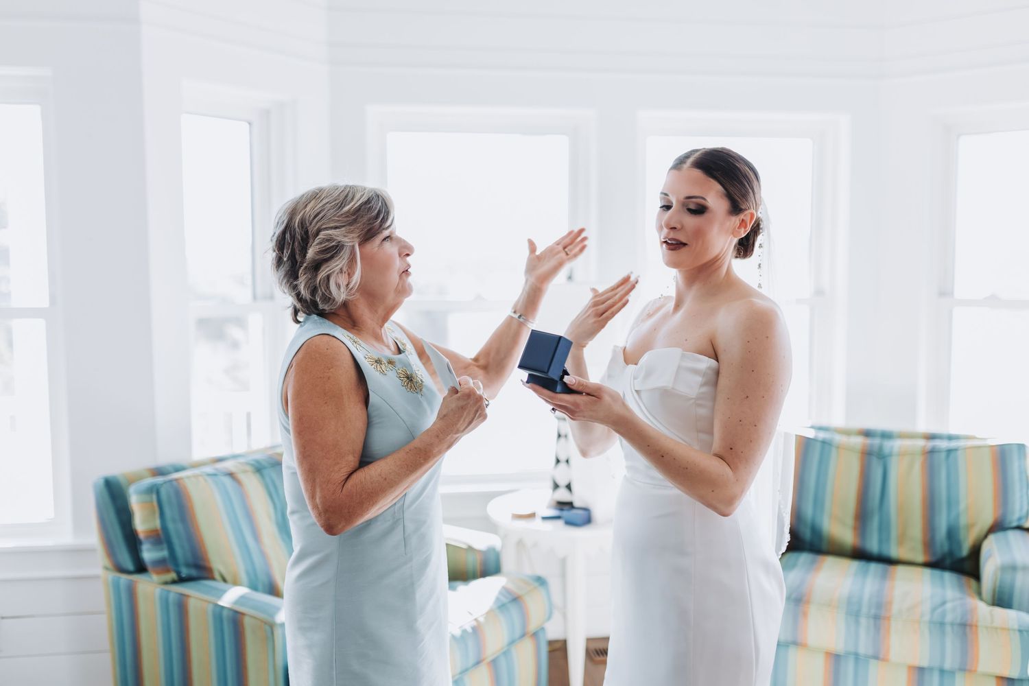 Bride in strapless wedding dress reacting with surprise to a gift from her mother in a bright room.