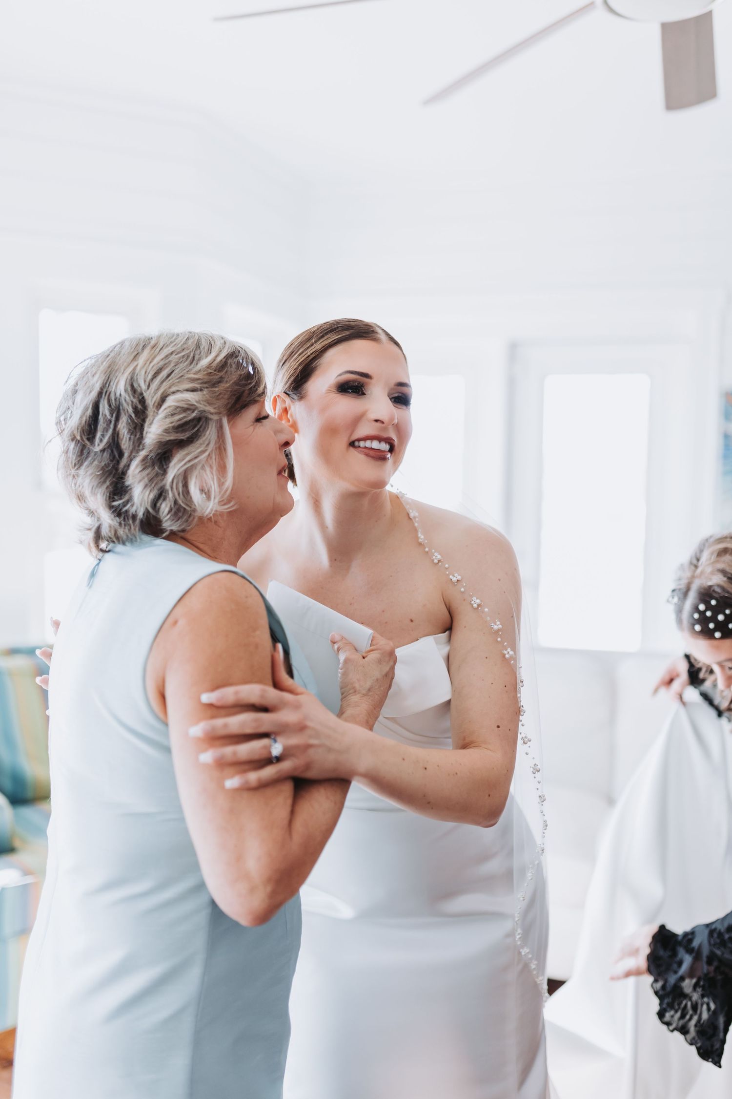 Bride in white strapless gown shares a joyful embrace with older woman in light blue dress indoors.