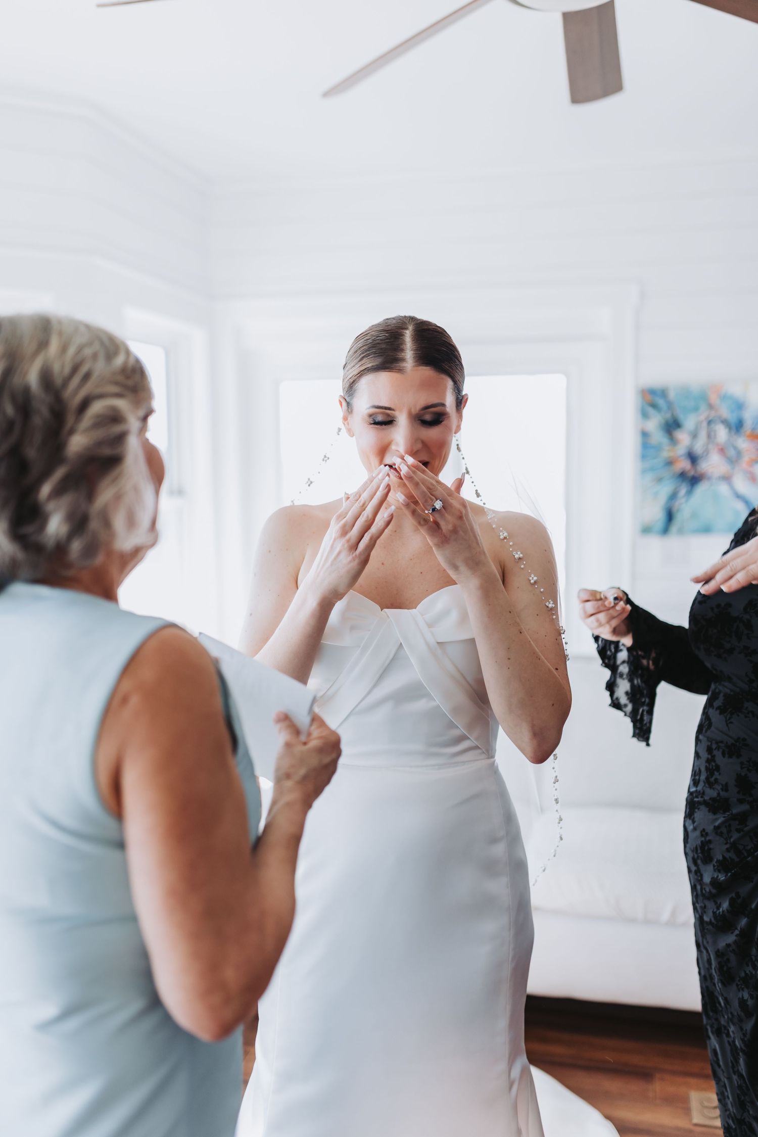 Emotional bride covers mouth with hands as family helps her into white wedding gown.