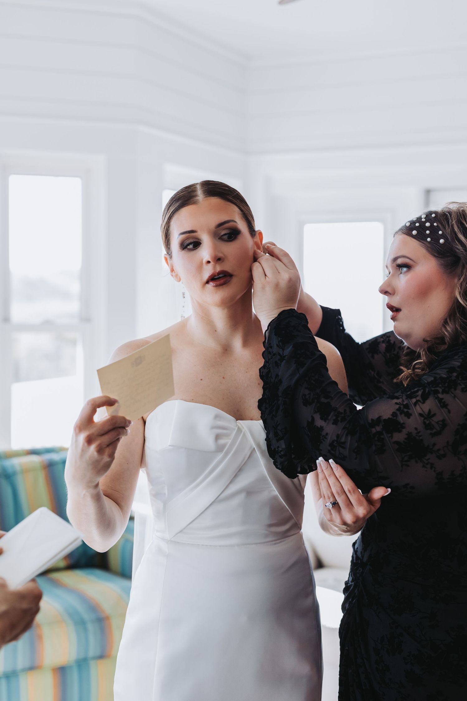 Bride in white strapless gown being helped with earrings while holding a letter before wedding.