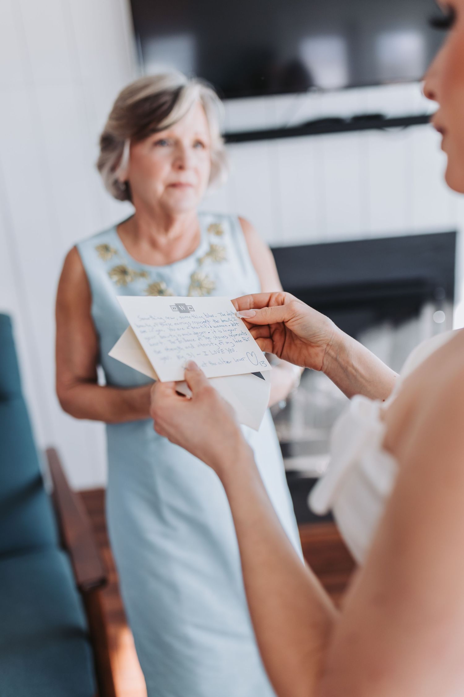 Bride reads a handwritten letter while older woman in light blue dress watches emotionally during wedding preparations.