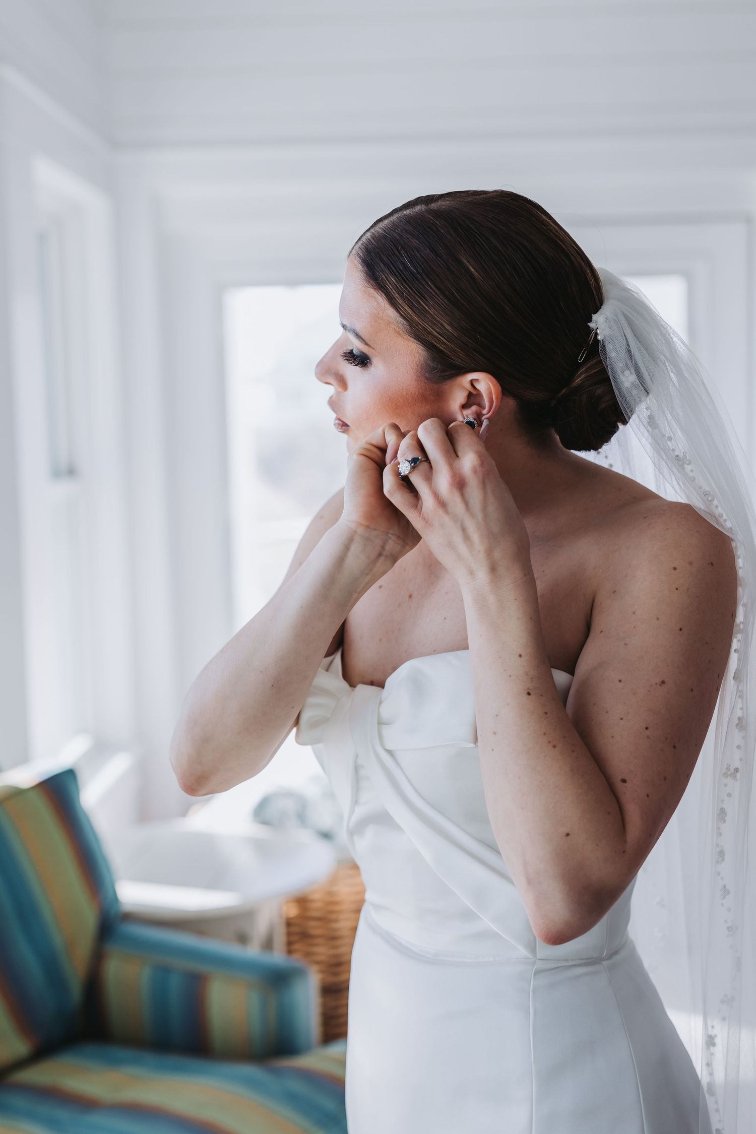 Bride in strapless white gown adjusting her earring while wearing a veil on her wedding day.